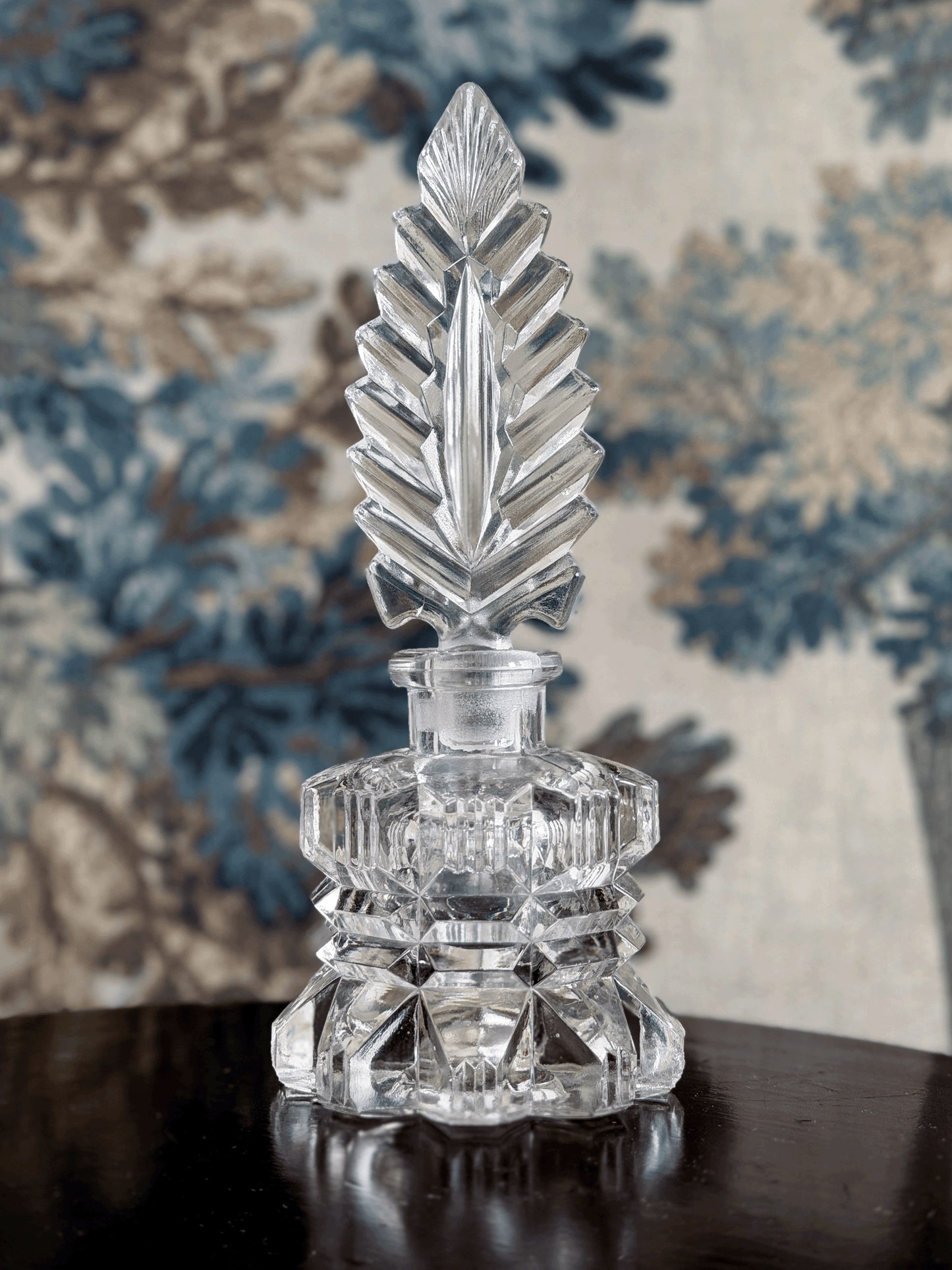 A clear cut-crystal perfume bottle with a faceted geometric feather stopper and a tiered base, displayed on a dark reflective surface against a blue and beige floral patterned fabric background.