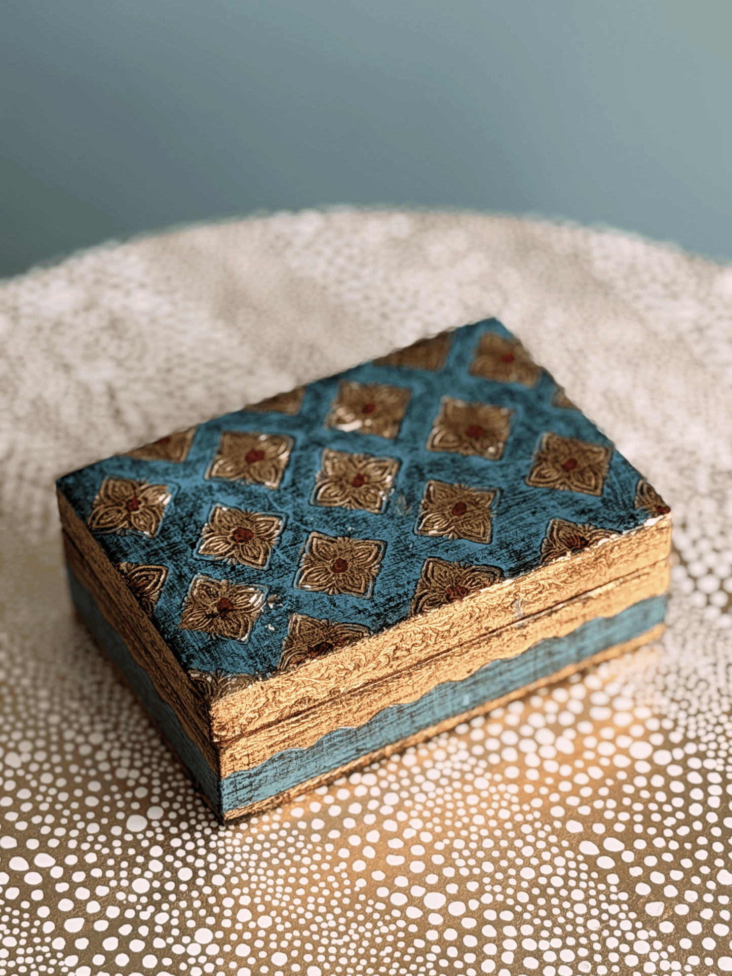 A small rectangular decorative box with a blue and gold hand-painted patterned lid, resting on a textured beige surface against a soft blue-gray background.