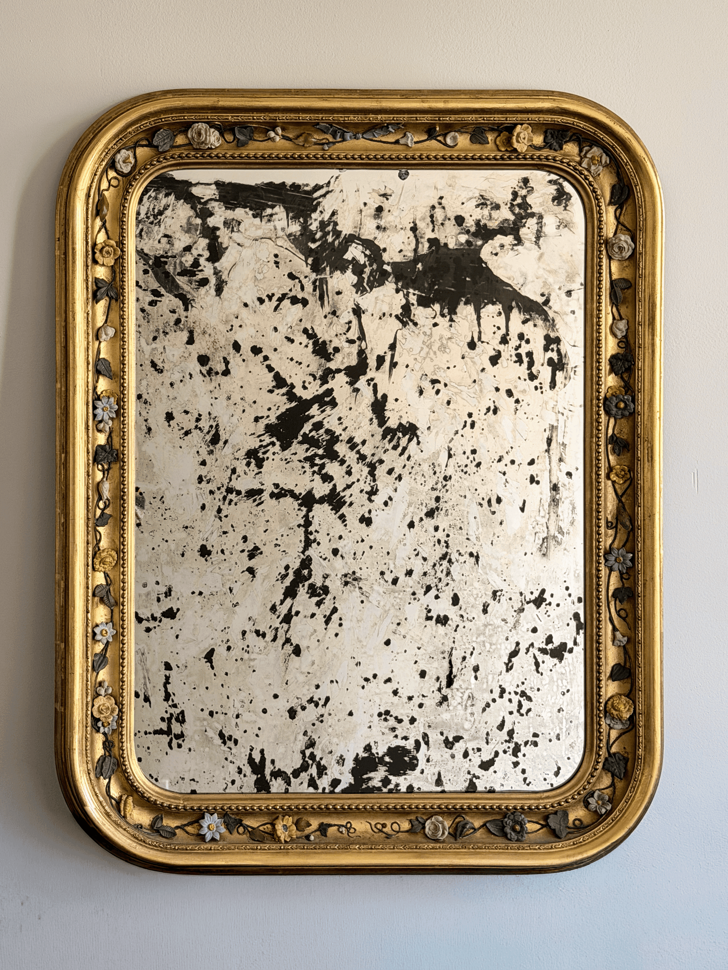 Front view of a 37 by 30 inch 19th-century French Louis Philippe mirror with a gold leaf giltwood frame, rounded top corners, an intricate porcelain floral vine border, and highly distressed, heavily foxed antiqued glass hanging on a light-colored wall.