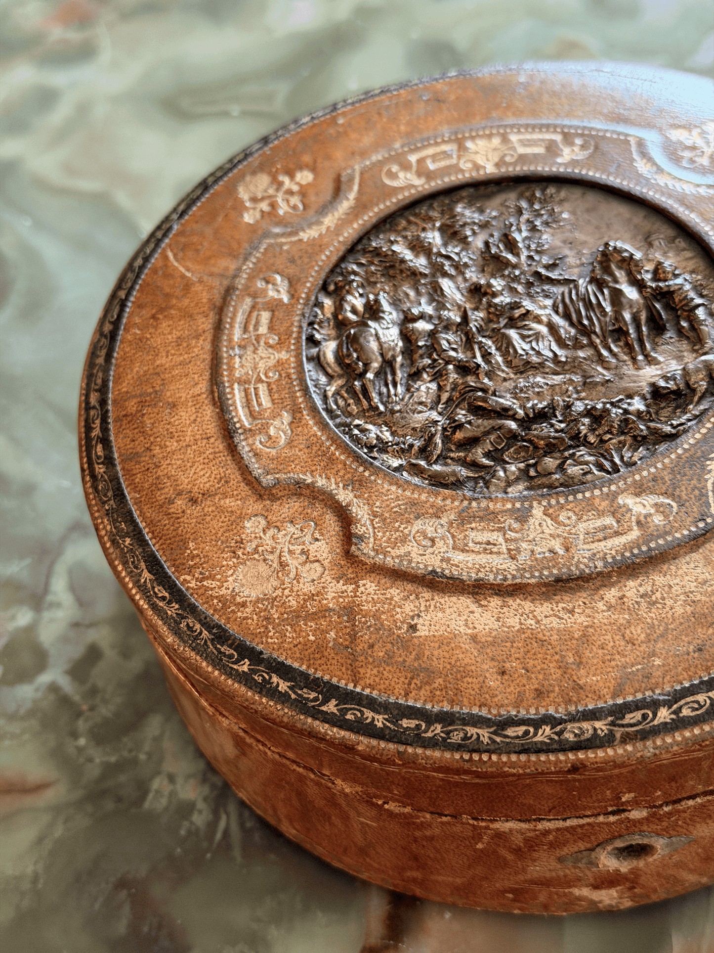 An antique round wooden box with a carved lid featuring a central relief scene and decorative floral patterns, resting on a mottled stone surface.