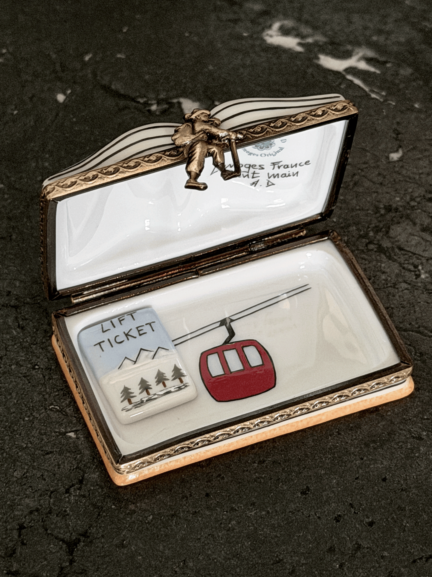 An open decorative trinket box with a white enamel interior and ornate gold-toned metal trim, featuring a small red cable car and a white lift ticket charm inside. The lid has a small figurine and handwritten text visible.