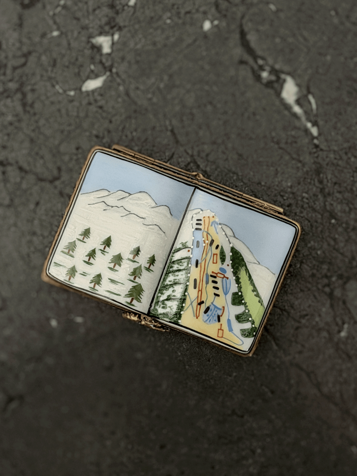 Hand-painted Rochard Limoges porcelain trinket box shaped like an open book, depicting a snow-capped alpine mountain peak on the left page and a colorful ski resort trail map with chairlift networks on the right page.