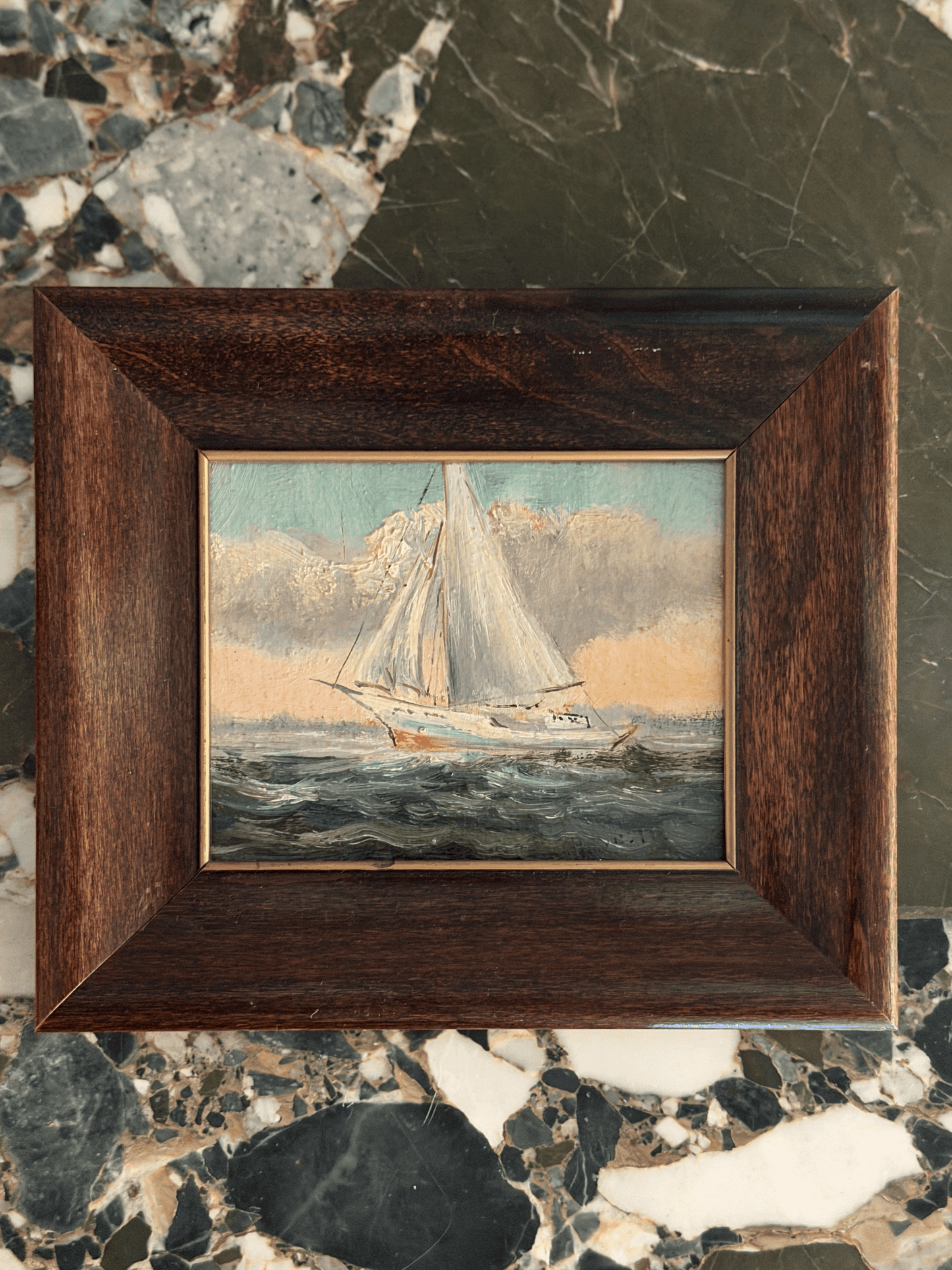 A framed miniature oil painting of a sailboat on the sea, set in a dark brown wooden frame and displayed on a polished marble surface.