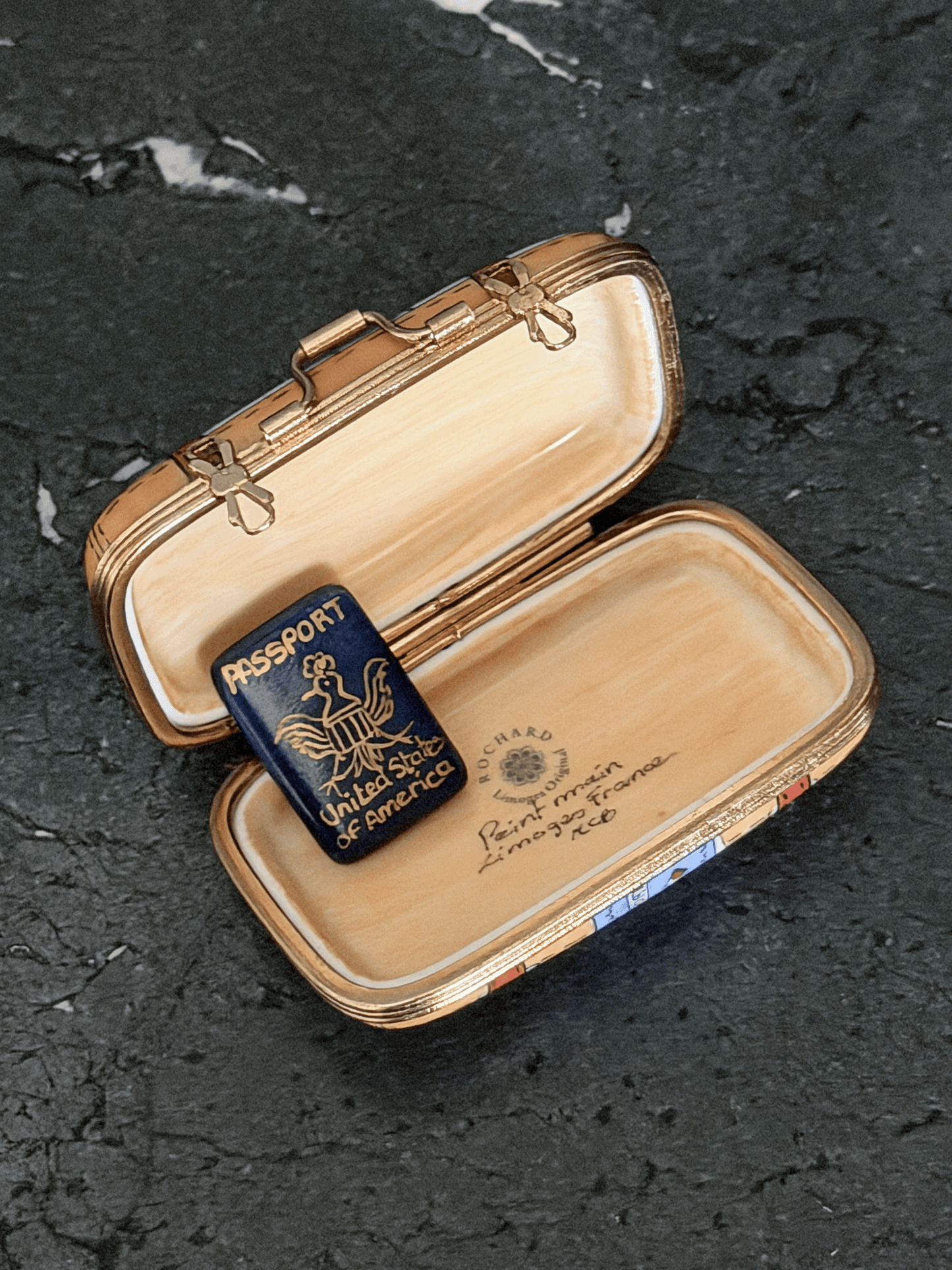 Opened interior of a Rochard map suitcase Limoges box showing a tan leather textile texture lining, the factory original workshop backstamp, and a separate loose removable miniature navy blue porcelain United States Passport accessory with a gold bald eagle seal.