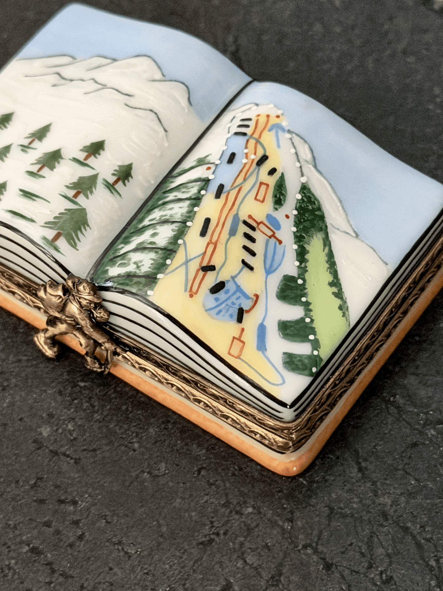 Hand-painted Rochard Limoges porcelain trinket box shaped like an open book, depicting a snow-capped alpine mountain peak on the left page and a colorful ski resort trail map with chairlift networks on the right page.