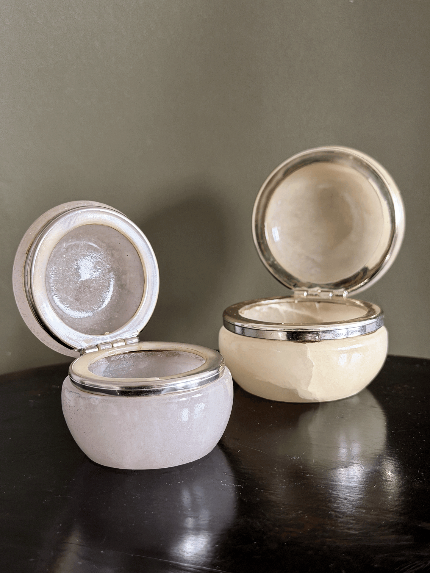 Two small round genuine Italian alabaster carved vanity jars, shown open on a dark reflective surface against a muted olive-gray background.