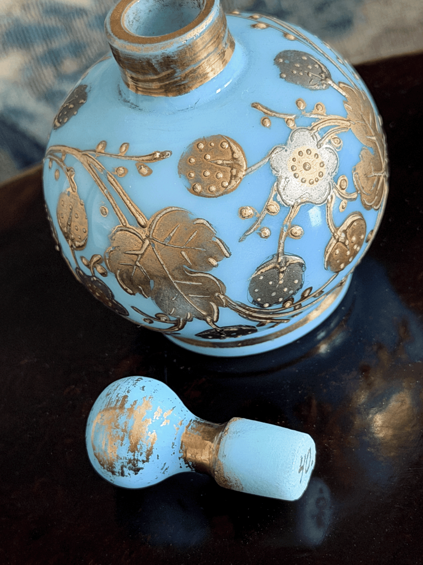 A light blue opaline glass perfume bottle with gold floral patterns, shown with its matching light blue and gold cap resting on a dark reflective surface.