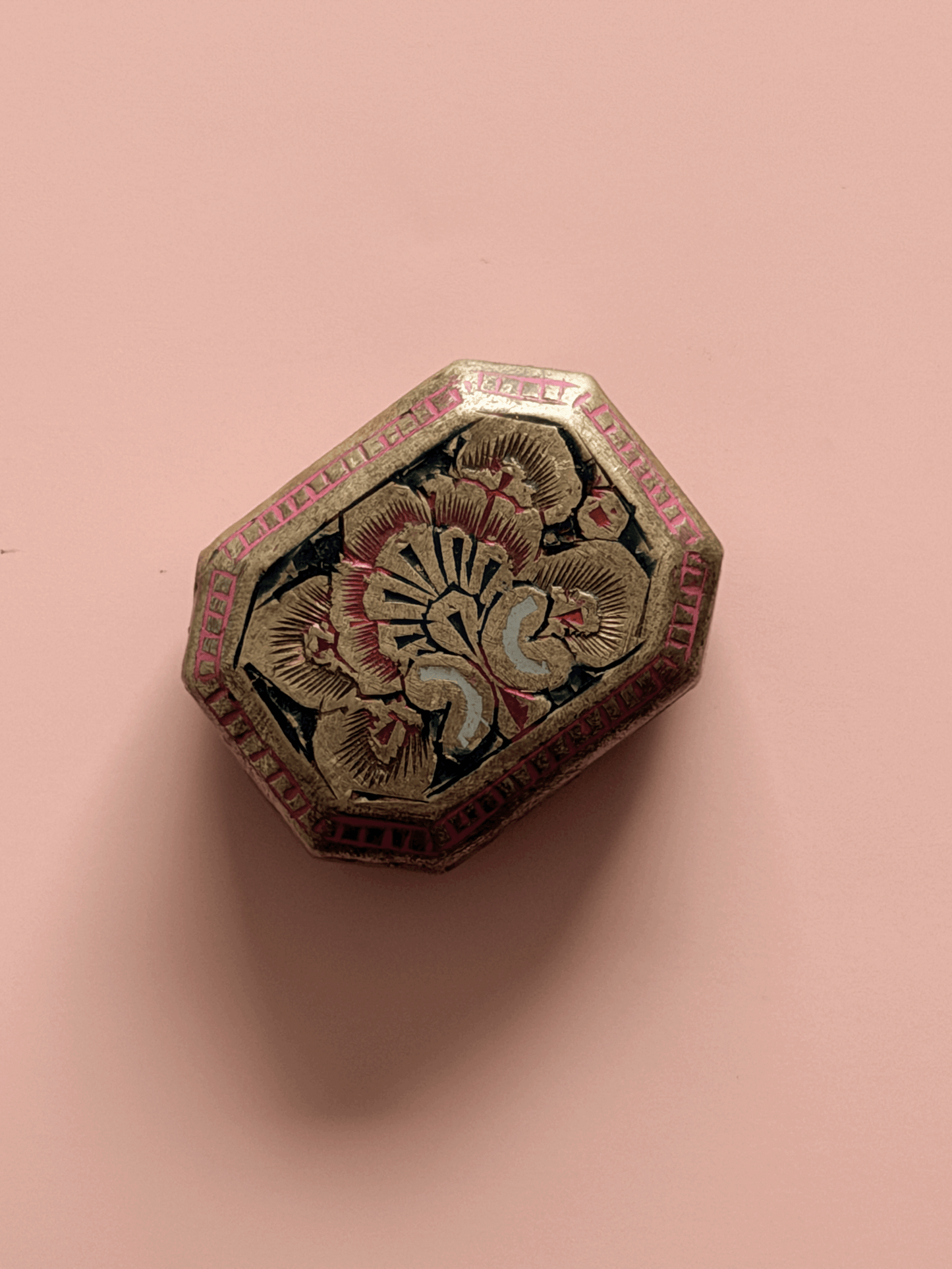 An overhead view of a small, antique octagonal brass trinket box against a pink background. The lid features an intricate, hand-engraved floral design filled with black enamel, surrounded by a border of bright pink enameled geometric cells.