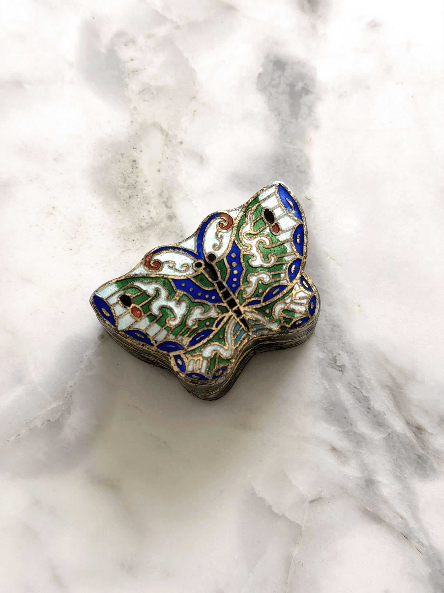 A small butterfly-shaped enamel trinket box with intricate blue, green, gold, and white detailing, resting on a white and gray marble surface.
