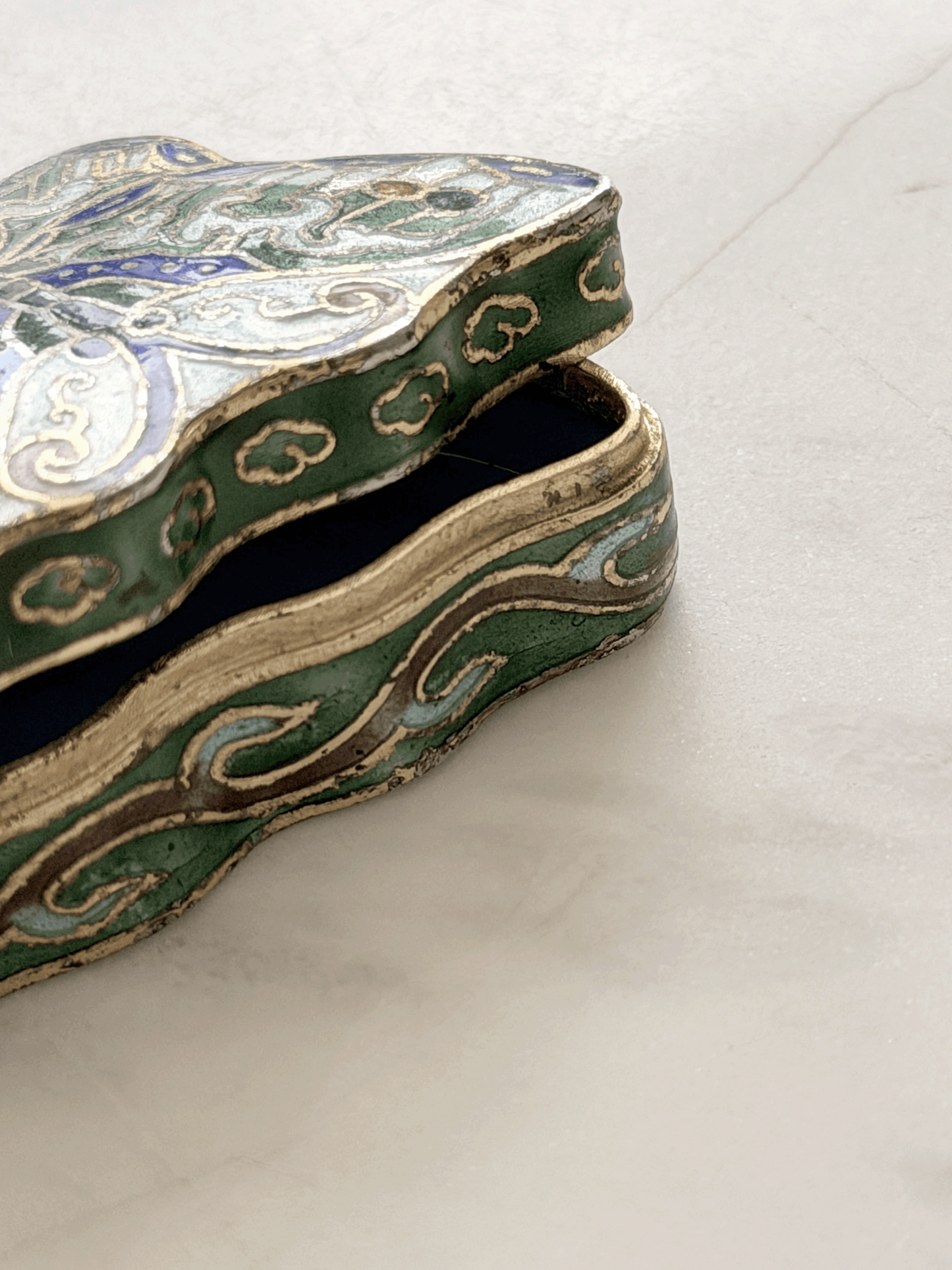 An ornate, tiny vintage butterfly-shaped trinket box with a green enamel body, gold-toned metal trim, and blue and white floral details, shown open on a light marble surface.