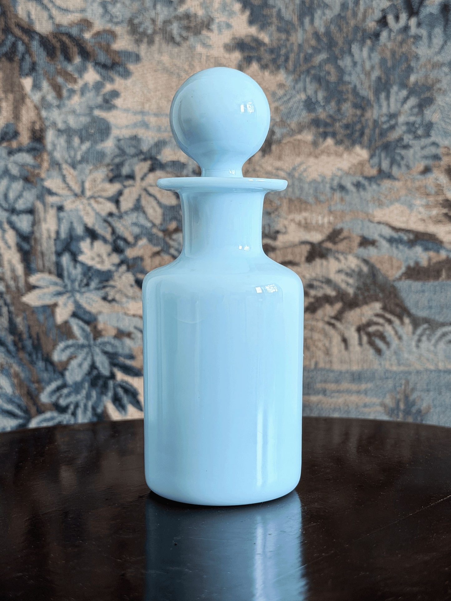 A light blue glass apothecary bottle with a matching round stopper, set on a dark reflective surface against a patterned blue and beige fabric backdrop.