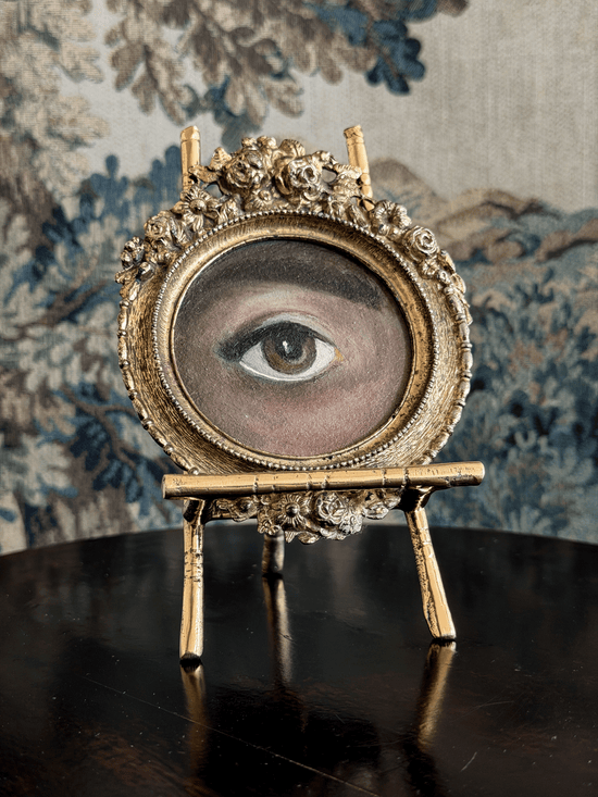 An ornate gold-framed oval tabletop mirror or picture frame featuring a painted human eye, displayed on a small gold stand over a dark reflective surface against a blue floral tapestry background.
