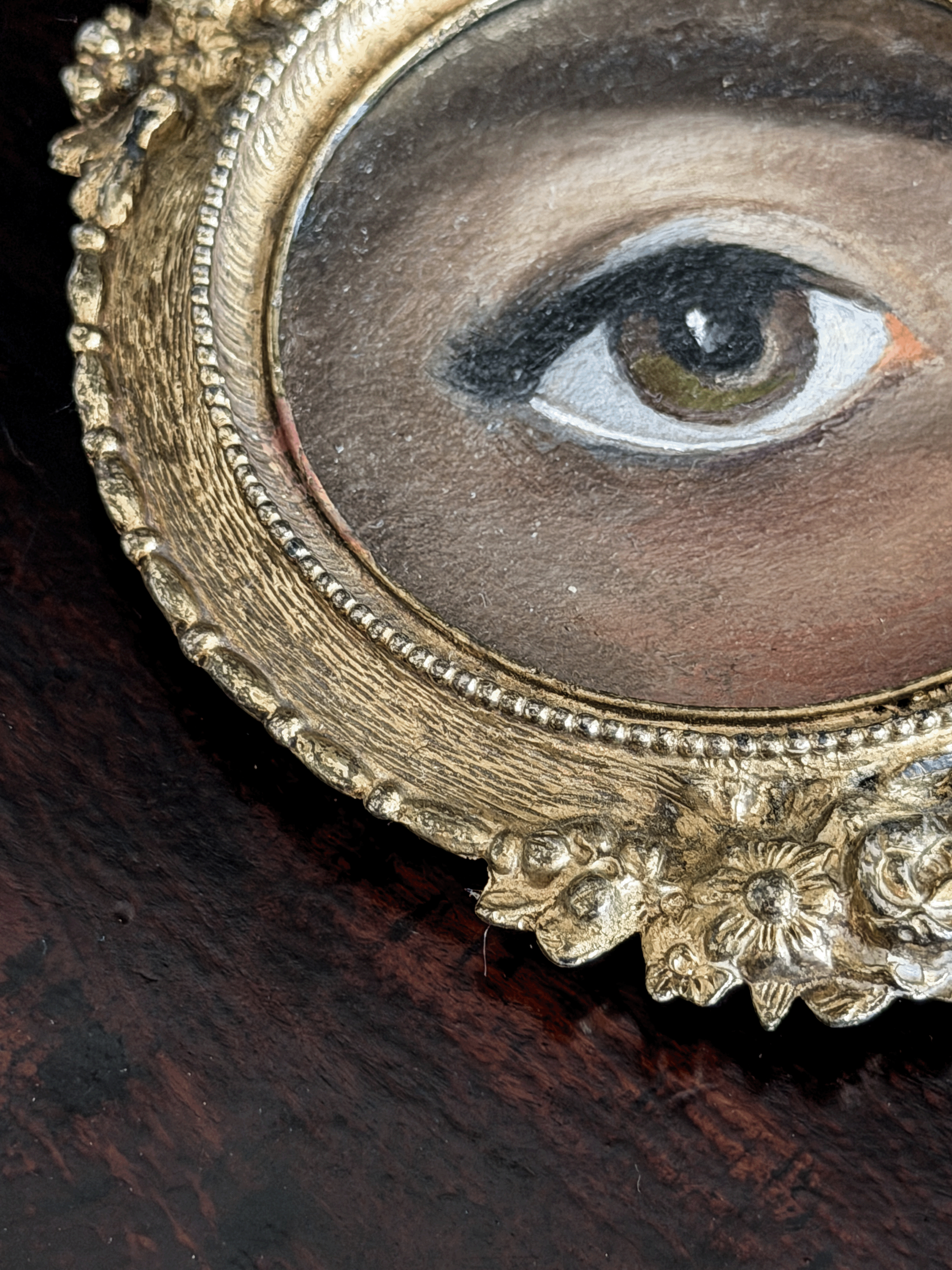 Close-up of an oval framed portrait painting showing a human eye, with an ornate gold-toned frame on a dark wooden surface.