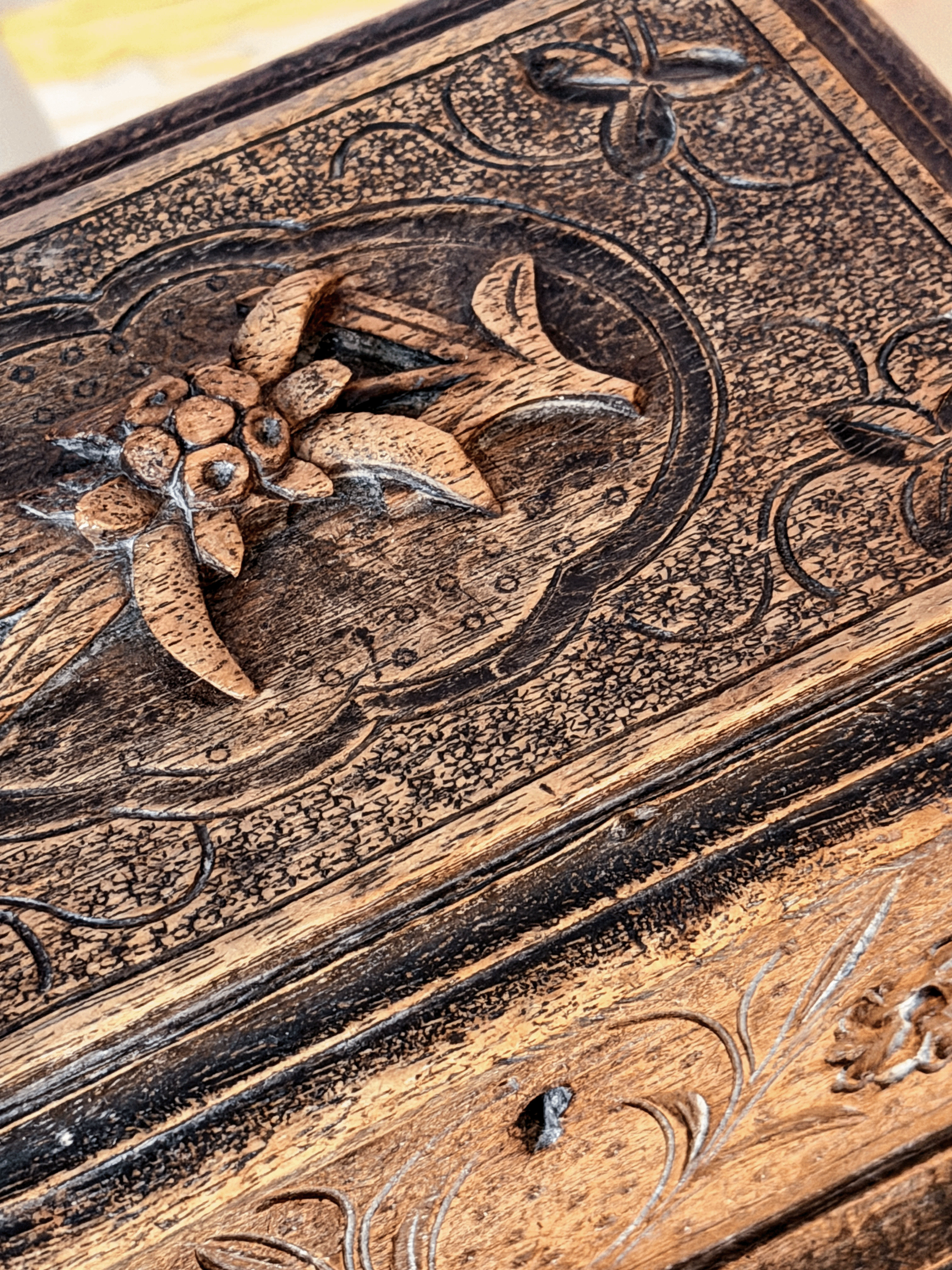 Close-up of a carved wooden box lid featuring floral and scroll motifs in dark brown and natural tan wood tones.