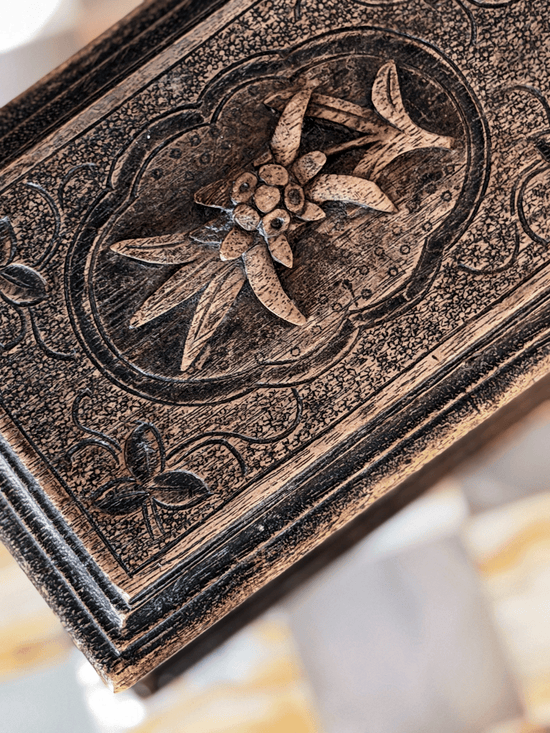 Close-up of the lid of an antique Brienz Black Forest style Swiss wood box, showcasing a high-relief hand-carved edelweiss mountain flower inside a scalloped frame against a hand-stippled background.