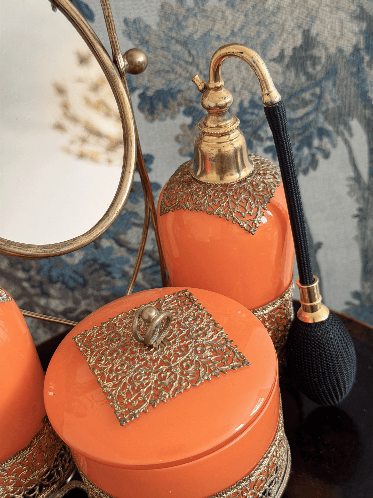 Orange bathroom vanity set with ornate gold-tone metal filagree, including a perfume atomizer, a round lidded powder/trinket jar with a decorative top piece in an intricate gold stand with integrated boudoir mirror.