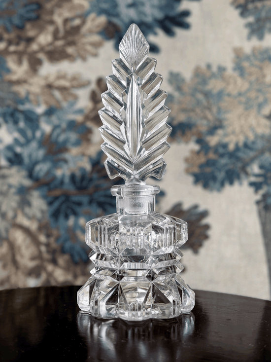 A clear cut-crystal perfume bottle with a faceted geometric feather stopper and a tiered base, displayed on a dark reflective surface against a blue and beige floral patterned fabric background.