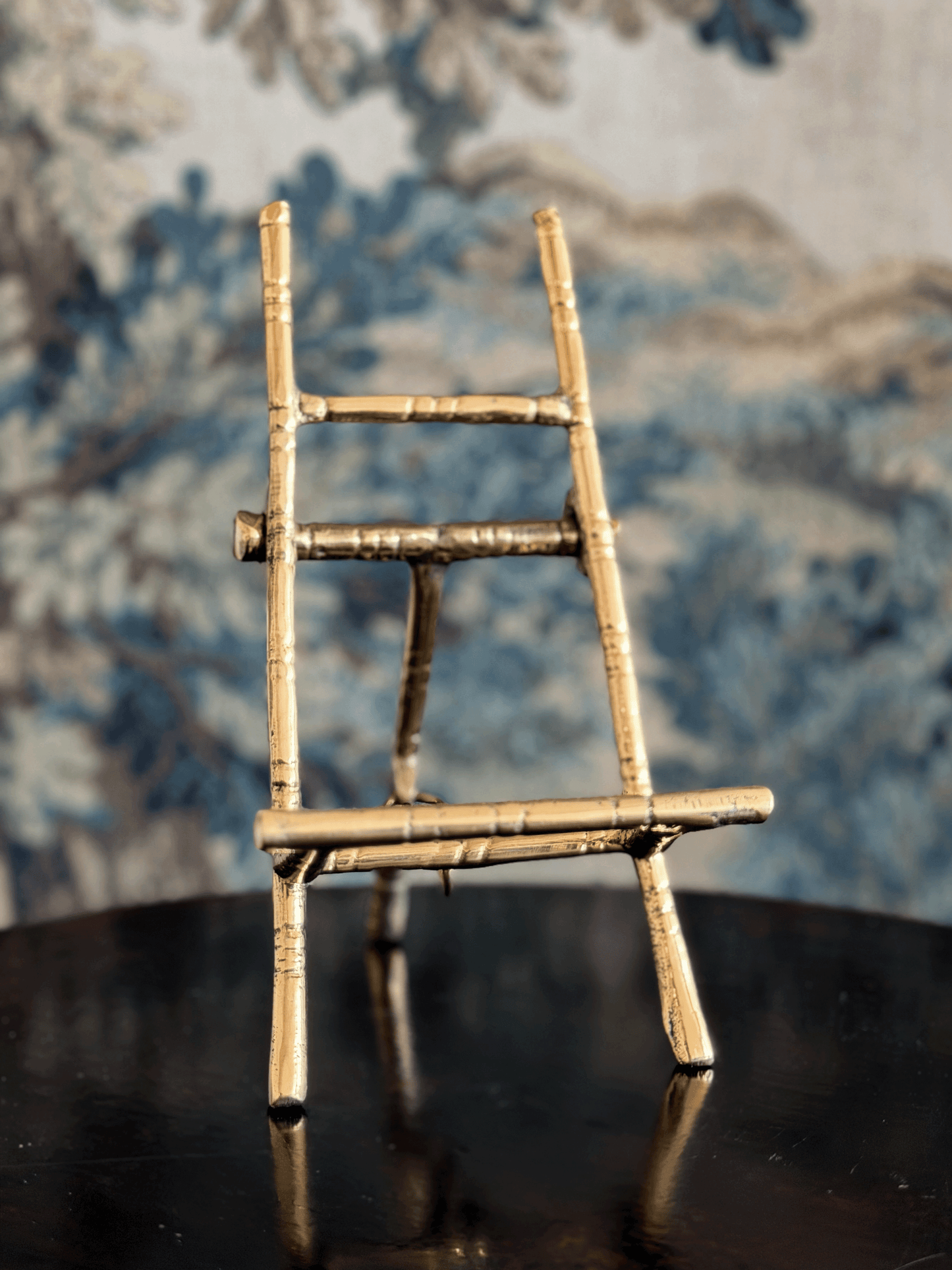 A miniature bamboo-style ladder stand with a natural light wood finish, displayed on a dark reflective surface against a blue and white patterned wallpaper background.