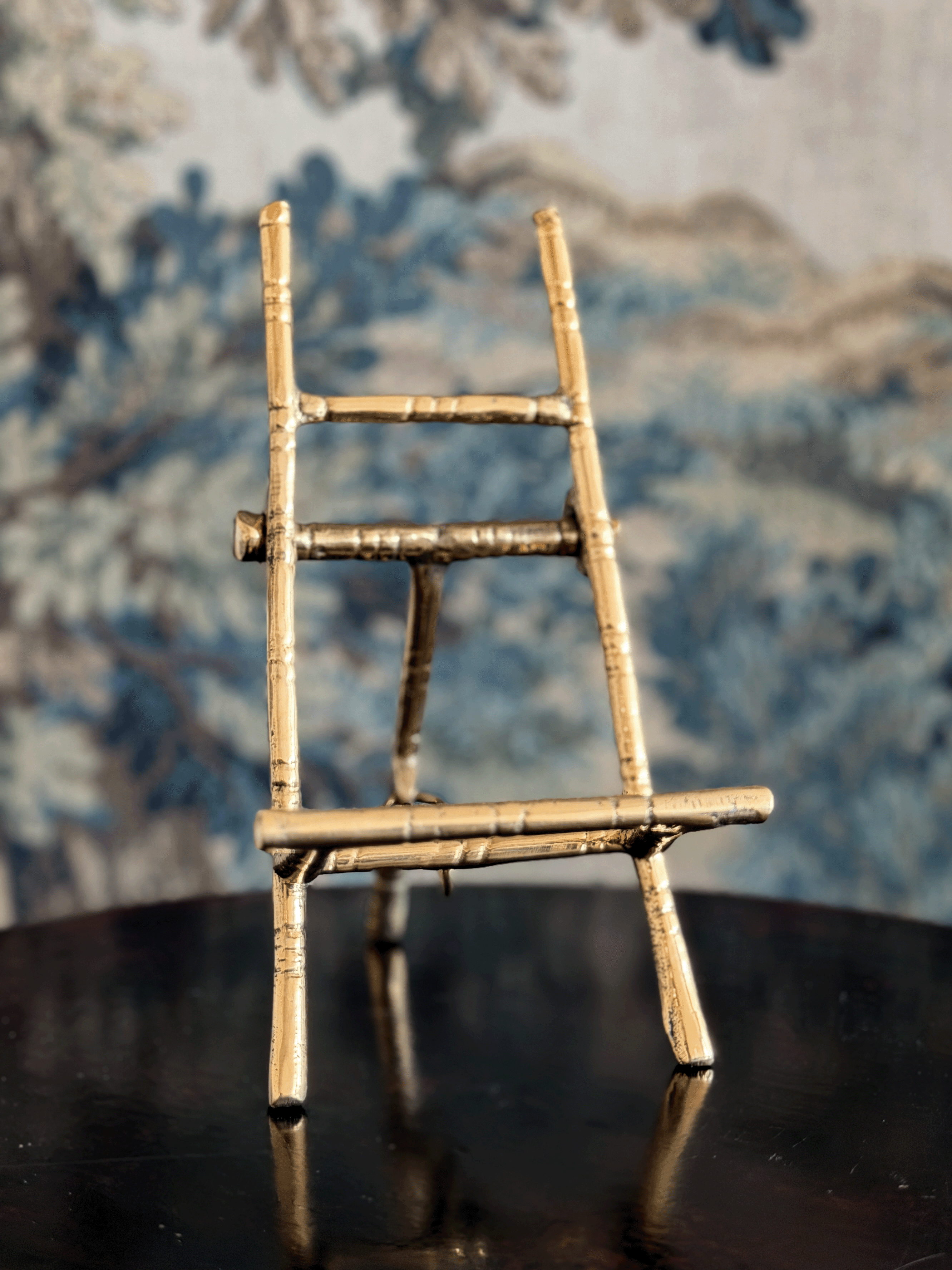 A miniature bamboo-style ladder stand with a natural light wood finish, displayed on a dark reflective surface against a blue and white patterned wallpaper background.