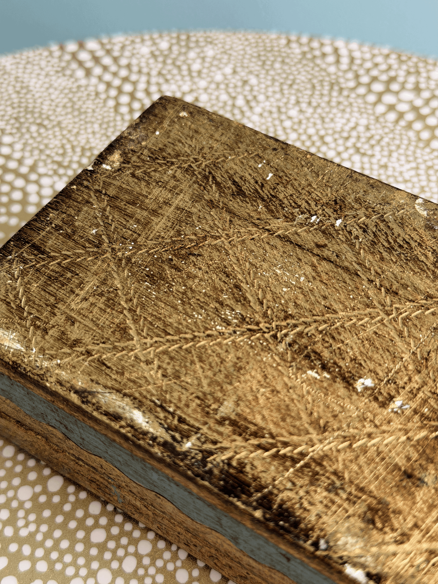 A close-up of a rectangular wooden box with a distressed, textured gold finish on top and light blue painted edges, resting on a patterned beige surface.