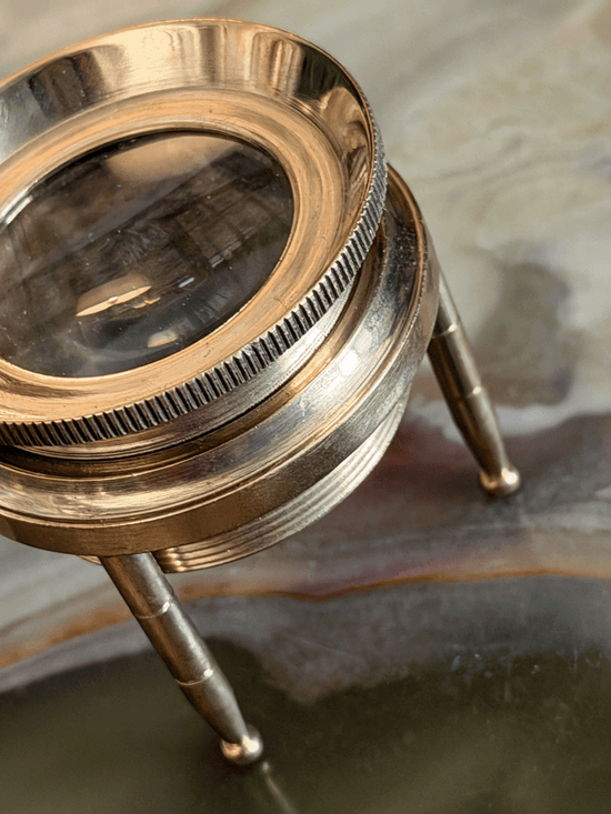 A close-up of a vintage brass magnifying glass stand with a clear glass lens and three legs, resting on a polished agate stone surface.