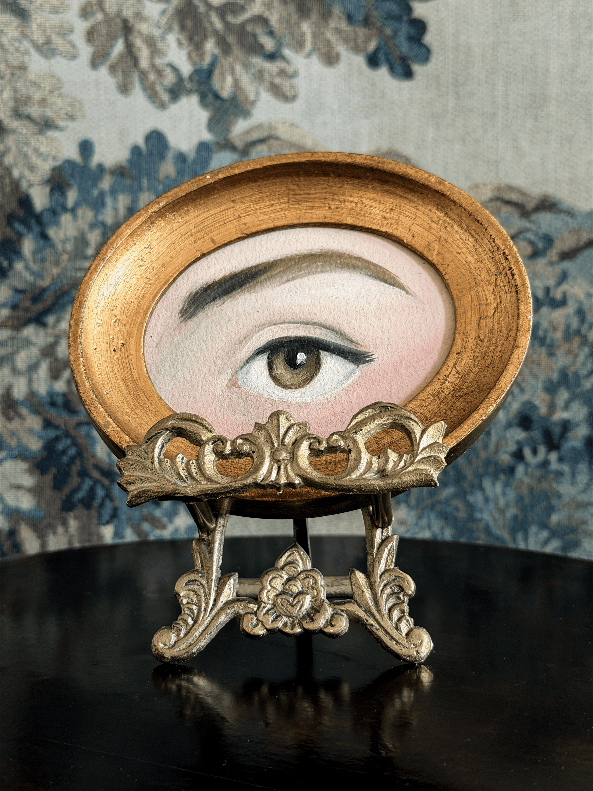 A decorative oval wall mirror or art piece featuring a painted human eye, displayed on an ornate gold stand against a blue floral patterned backdrop.