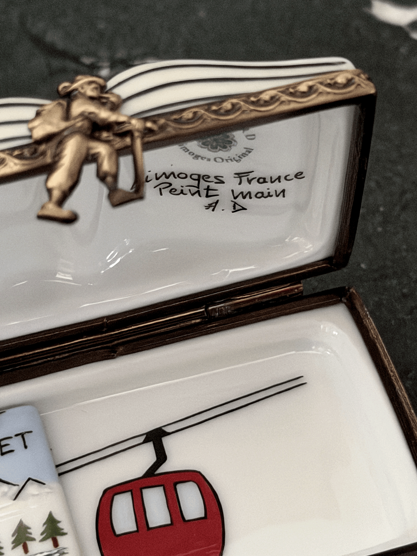 Interior view of an opened Rochard Limoges ski book box showing a hand-painted red gondola car, an authenticated Limoges France backstamp, a sculpted skier figurine brass clasp, and a loose removable miniature porcelain lift ticket insert.