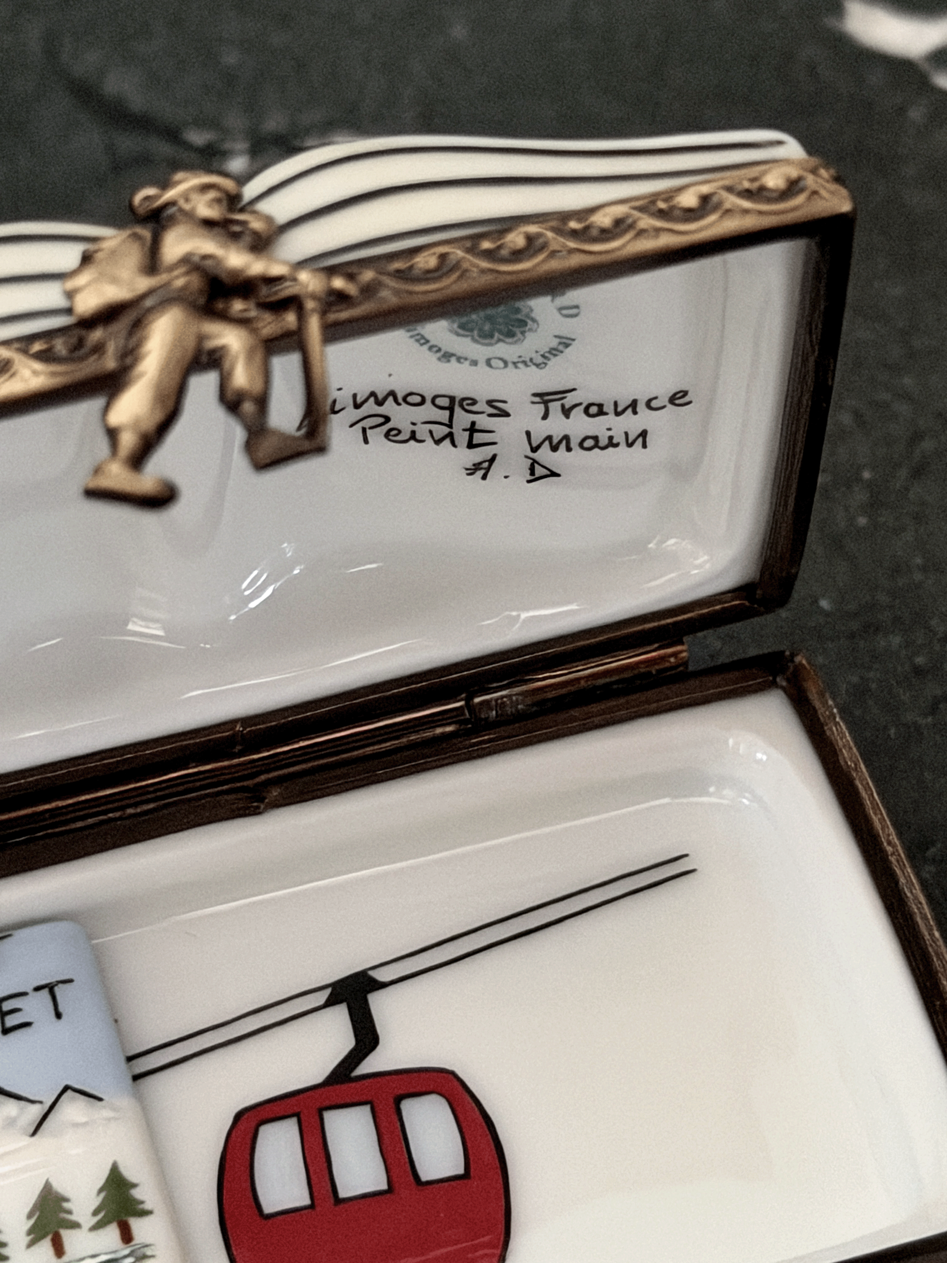 Interior view of an opened Rochard Limoges ski book box showing a hand-painted red gondola car, an authenticated Limoges France backstamp, a sculpted skier figurine brass clasp, and a loose removable miniature porcelain lift ticket insert.