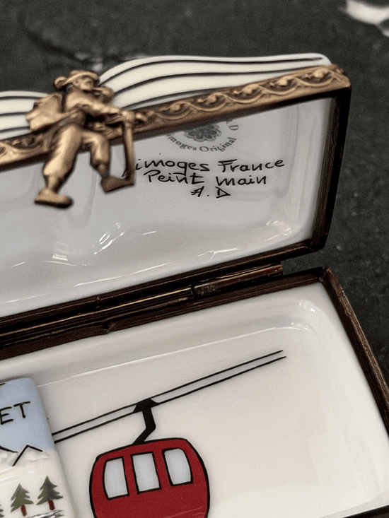 Interior view of an opened Rochard Limoges ski book box showing a hand-painted red gondola car, an authenticated Limoges France backstamp, a sculpted skier figurine brass clasp, and a loose removable miniature porcelain lift ticket insert.
