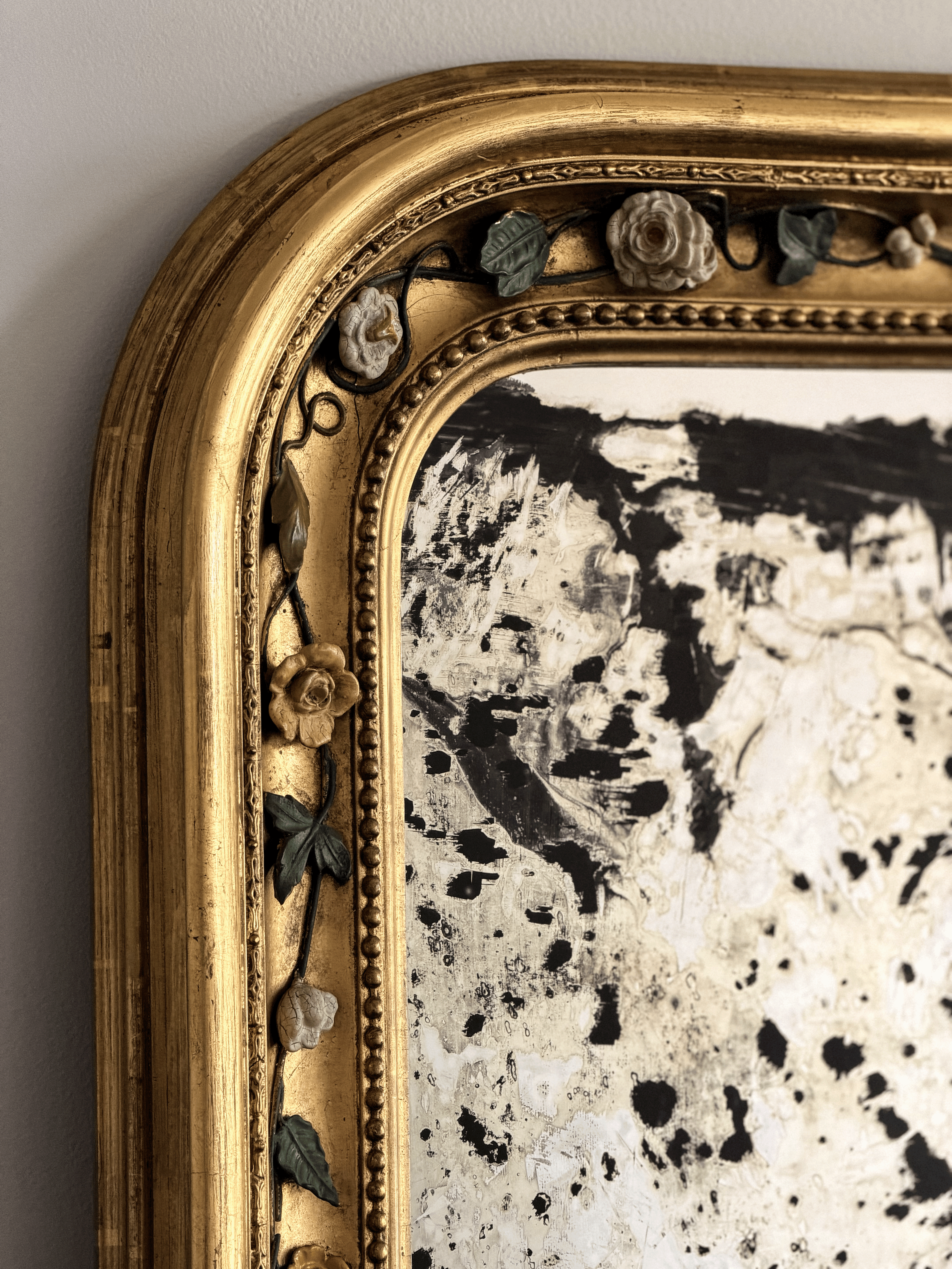Detailed close-up of a 19th-century French mirror frame showcasing hand-modeled porcelain white and yellow roses attached to hand-wrought dark metal vine moldings against a brilliant gold leaf gilded finish, next to a heavily foxed antiqued glass mirror plate.