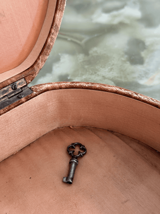 A small antique-style metal key with a decorative floral bow lies inside an open vintage-style trunk or box lined with tan fabric. 