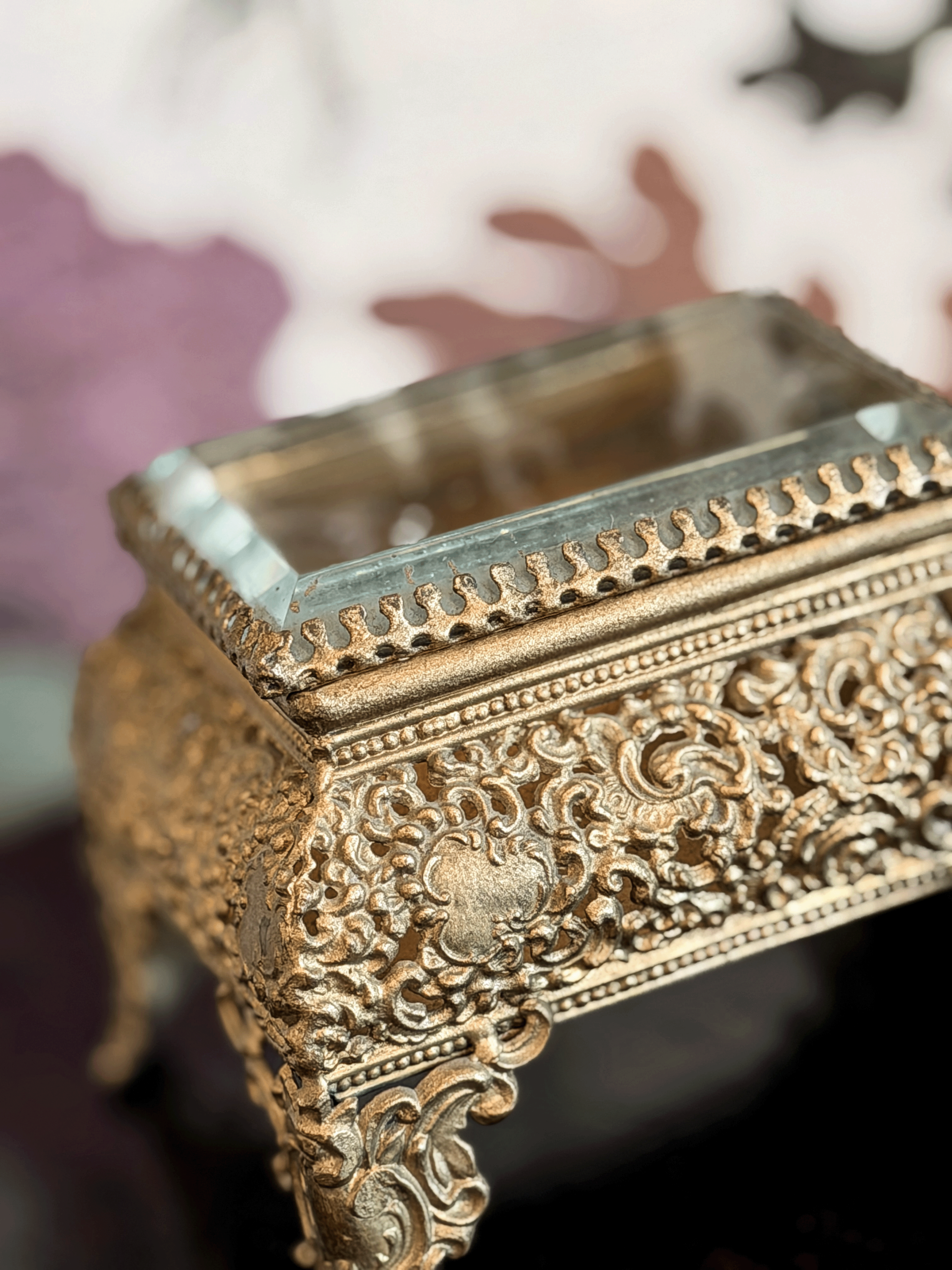 An ornate gold-toned metal decorative box with intricate embossed scrollwork and a clear glass top, shown in a close-up shot against a softly blurred background.