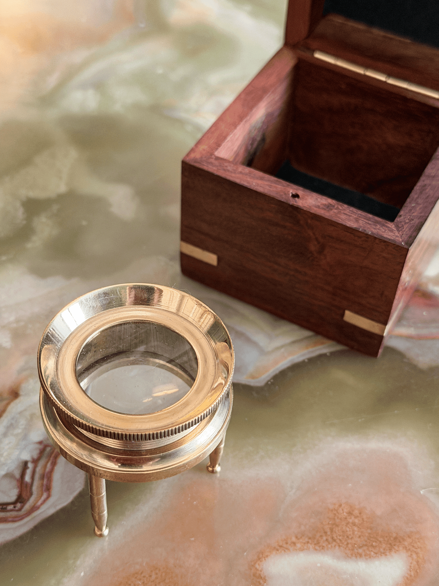 A brass magnifying glass on a small stand next to an open dark wood presentation box with brass corner accents, set on a polished marble surface.