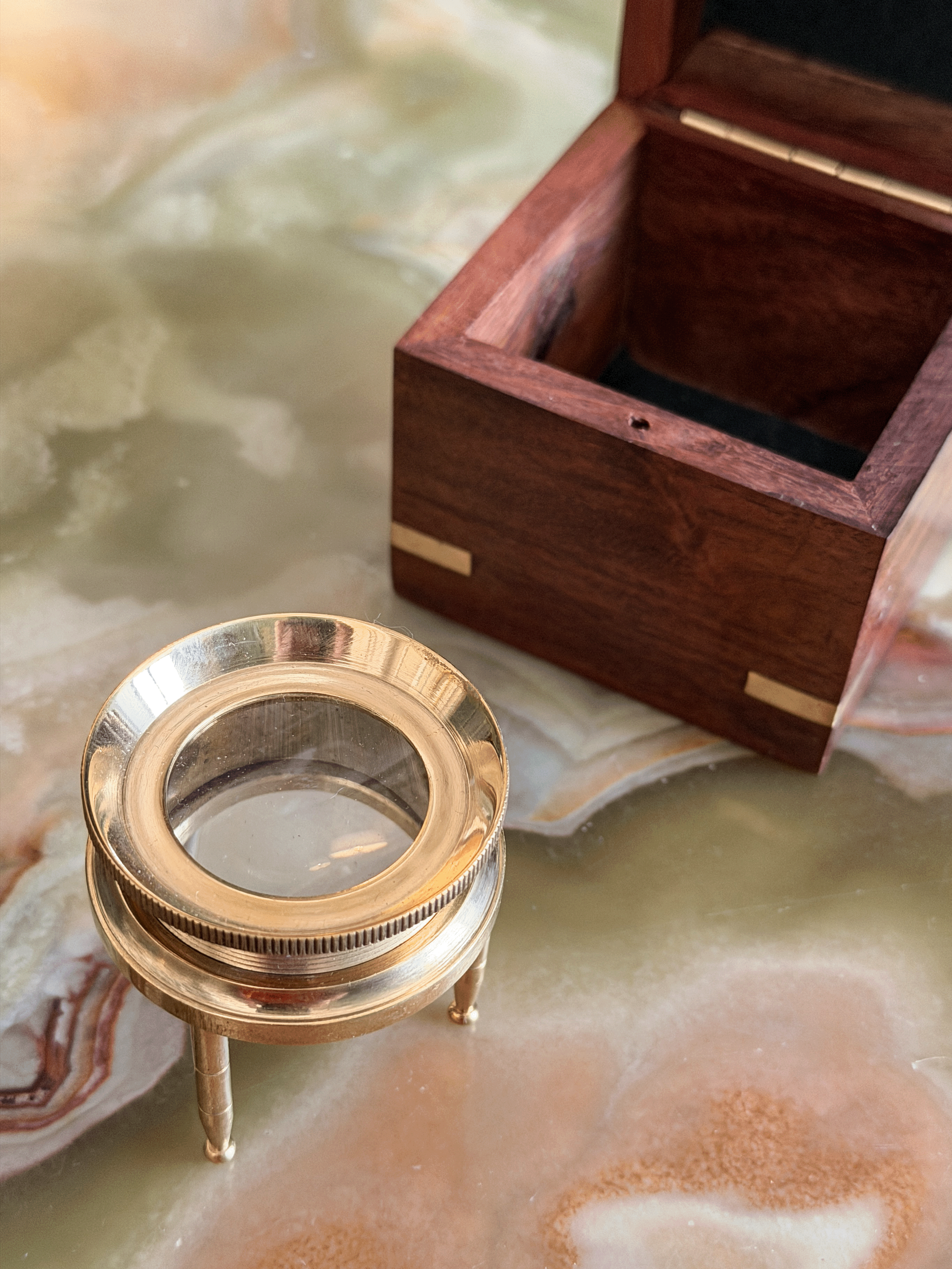 A brass magnifying glass on a small stand next to an open dark wood presentation box with brass corner accents, set on a polished marble surface.