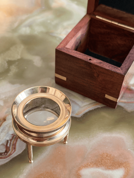 A brass magnifying glass on a small stand next to an open dark wood presentation box with brass corner accents, set on a polished marble surface.