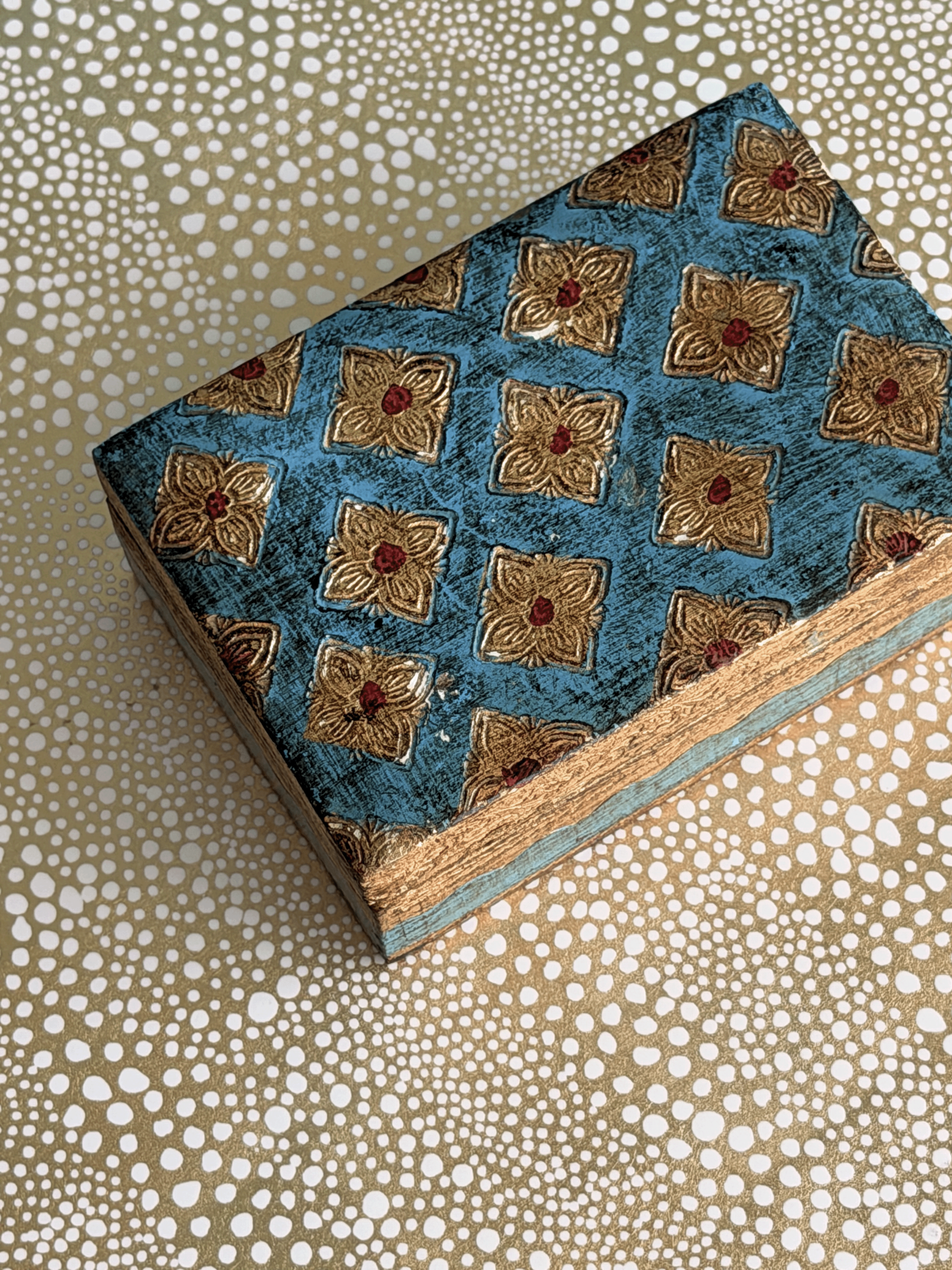 A rectangular decorative box with a distressed teal-blue lid featuring a repeating gold floral tile pattern and small red centers.