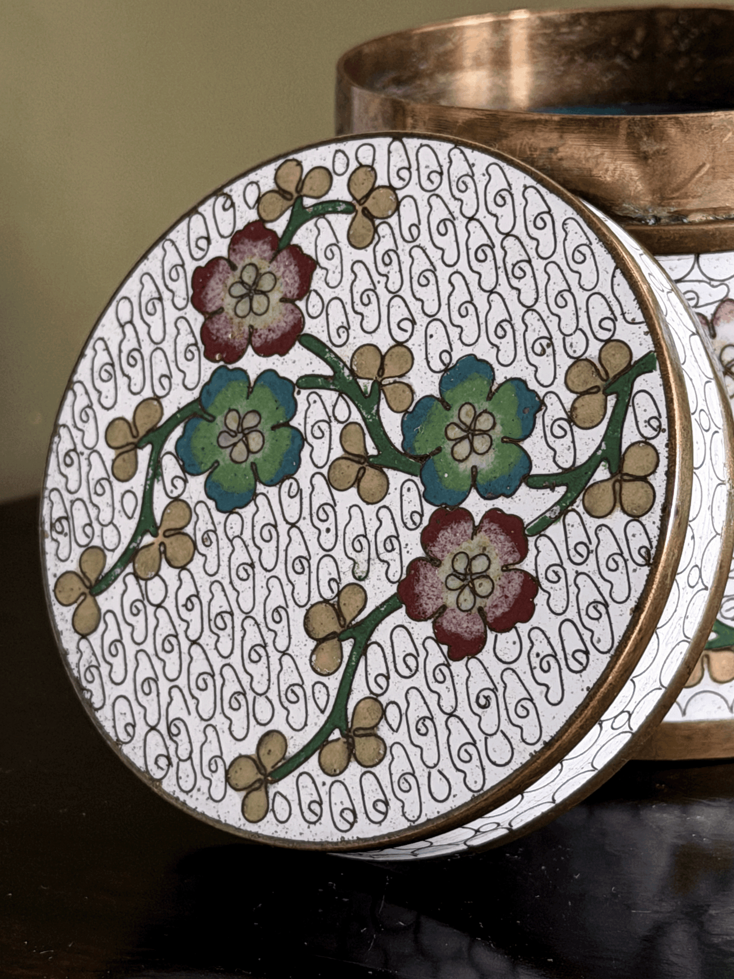 A close-up of a round decorative tin or container lid with a white background, black swirling line pattern, and floral motifs in green, blue, and burgundy, with a metallic bronze rim. A matching bronze container is partially visible behind it.