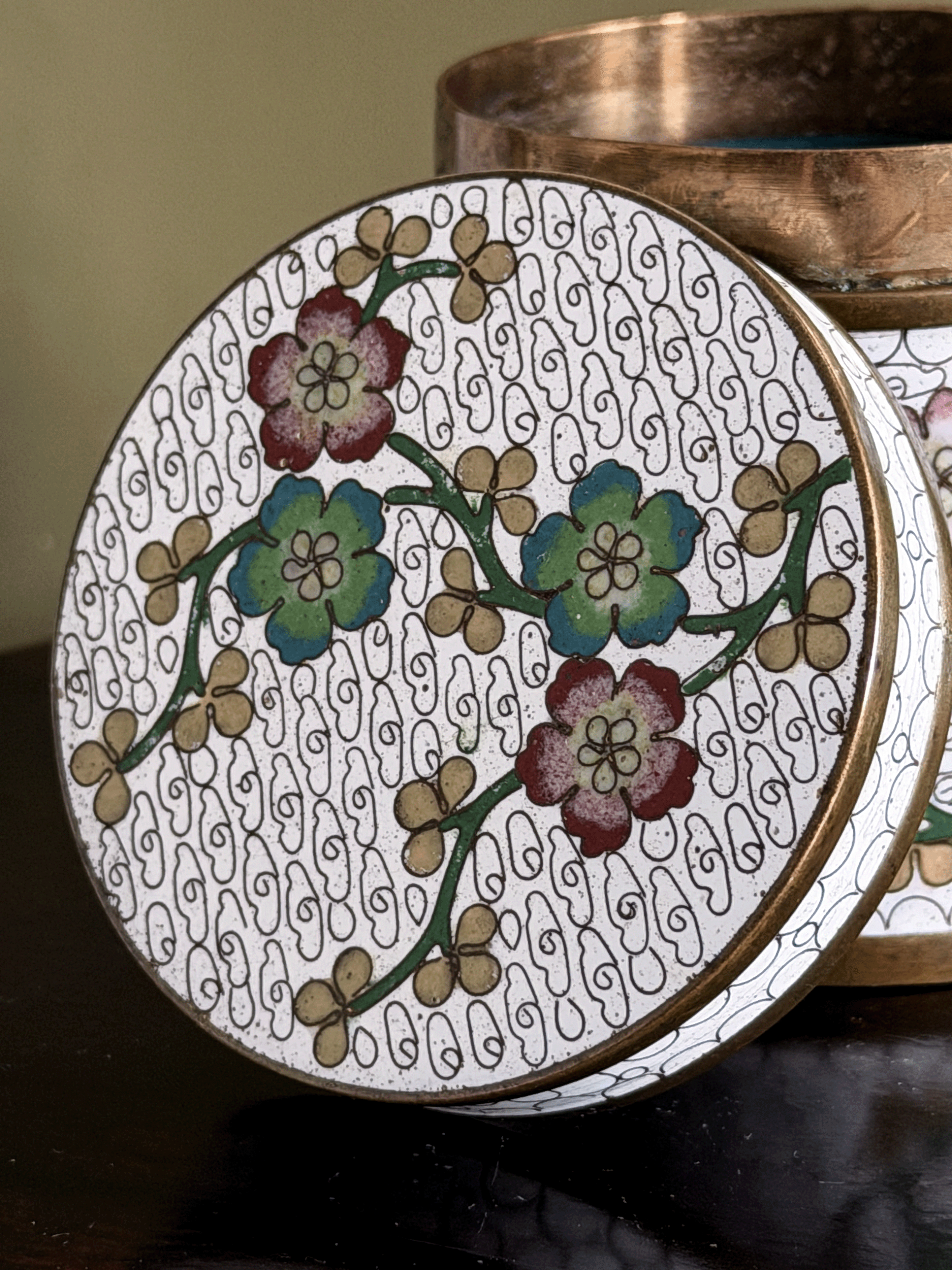 A close-up of a round decorative tin or container lid with a white background, black swirling line pattern, and floral motifs in green, blue, and burgundy, with a metallic bronze rim. A matching bronze container is partially visible behind it.