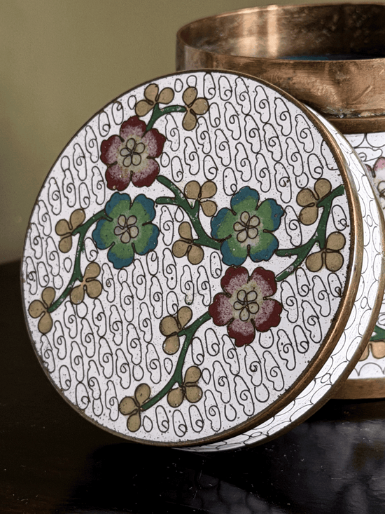 A close-up of a round decorative tin or container lid with a white background, black swirling line pattern, and floral motifs in green, blue, and burgundy, with a metallic bronze rim. A matching bronze container is partially visible behind it.