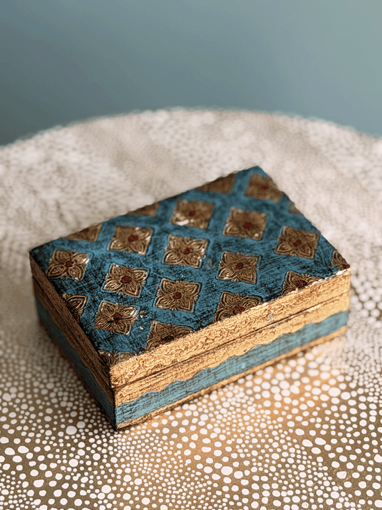 A small rectangular decorative box with a blue and gold hand-painted patterned lid, resting on a textured beige surface against a soft blue-gray background.