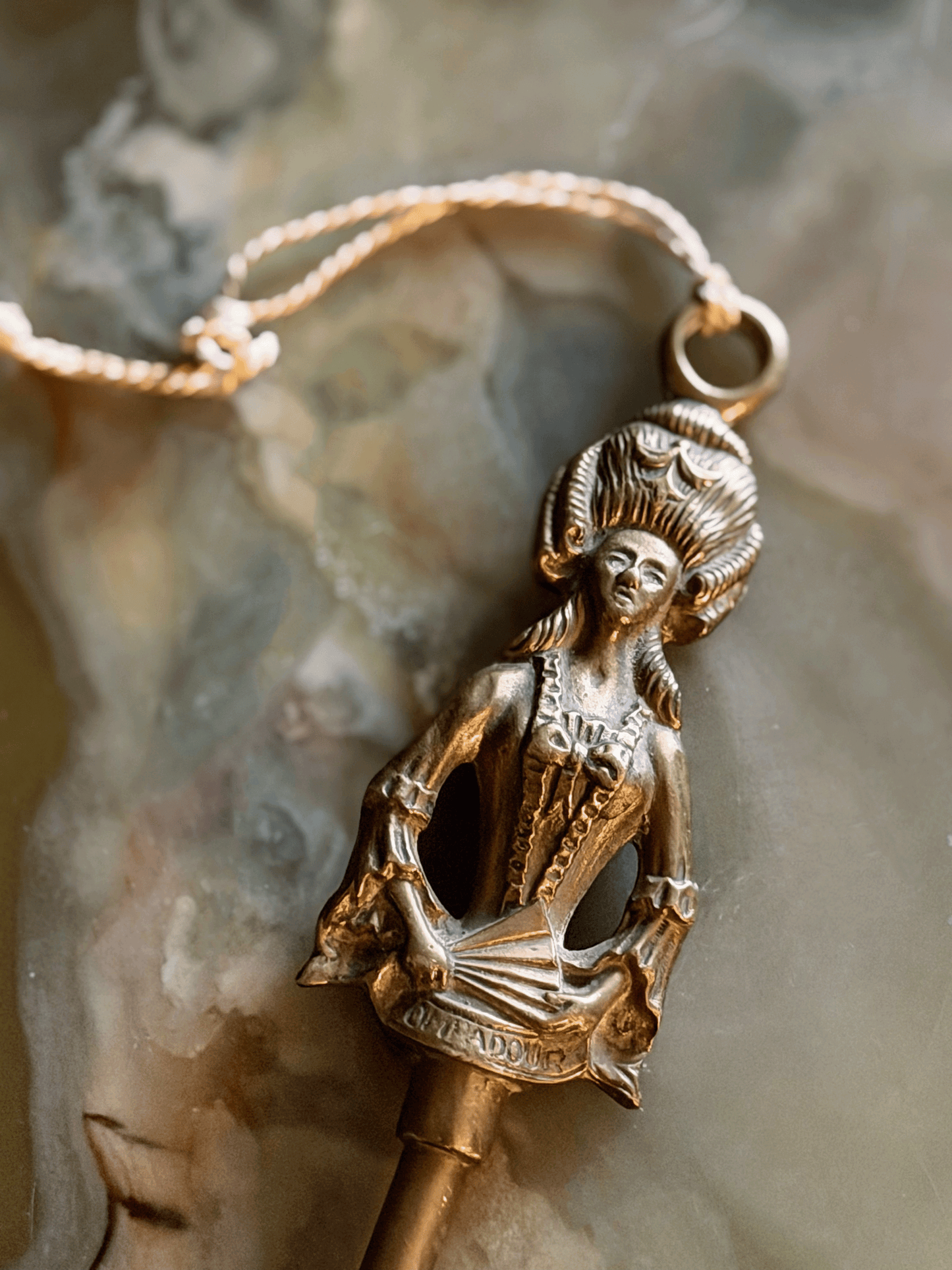 A detailed close-up view of the solid brass handle finial of a vintage fireplace toasting fork, resting on a swirling cream and green onyx surface. The finial is sculpted into the shape of Madame de Pompadour in 18th-century French dress with a towering, high-swept hairstyle and a laced bodice. She holds a folded hand fan across her front that has the word POMPADOUR stamped clearly into it. A thin, braided cream string tassel is tied to a suspension ring at the very top of her head.