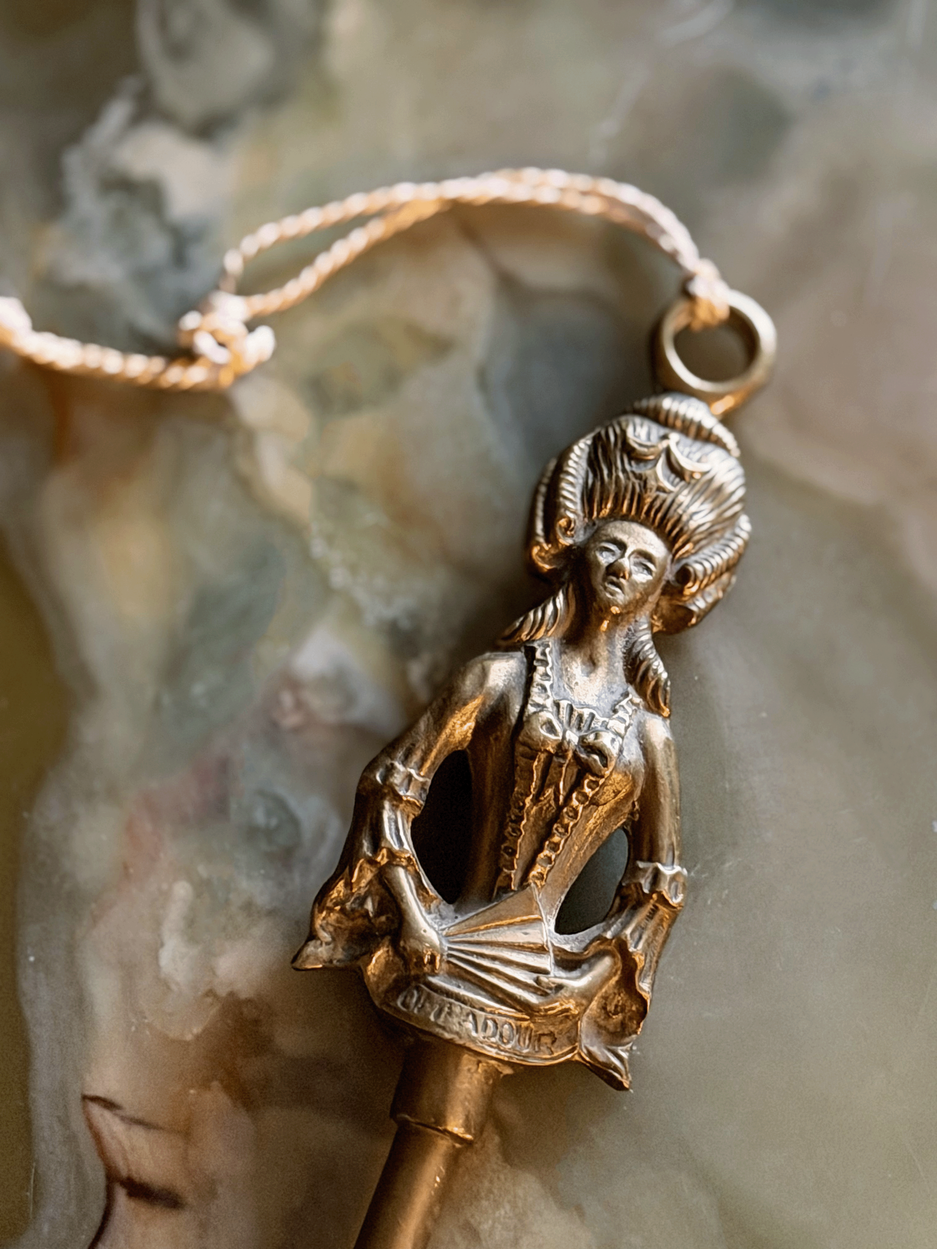 A detailed close-up view of the solid brass handle finial of a vintage fireplace toasting fork, resting on a swirling cream and green onyx surface. The finial is sculpted into the shape of Madame de Pompadour in 18th-century French dress with a towering, high-swept hairstyle and a laced bodice. She holds a folded hand fan across her front that has the word POMPADOUR stamped clearly into it. A thin, braided cream string tassel is tied to a suspension ring at the very top of her head.