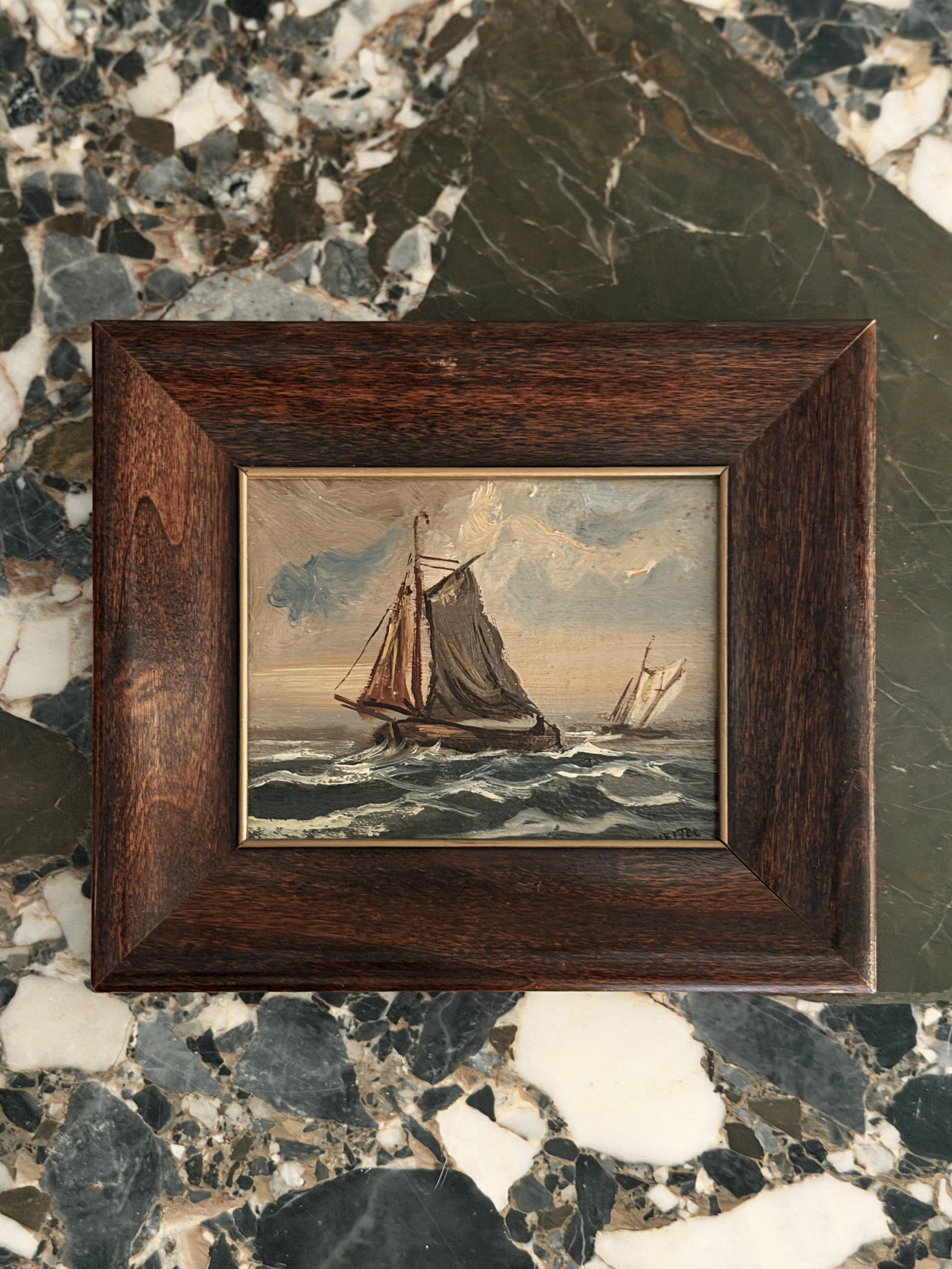 A small framed oil painting depicting two sailing ships on the ocean, set in a dark brown wooden frame and placed on a polished black and white marble surface.