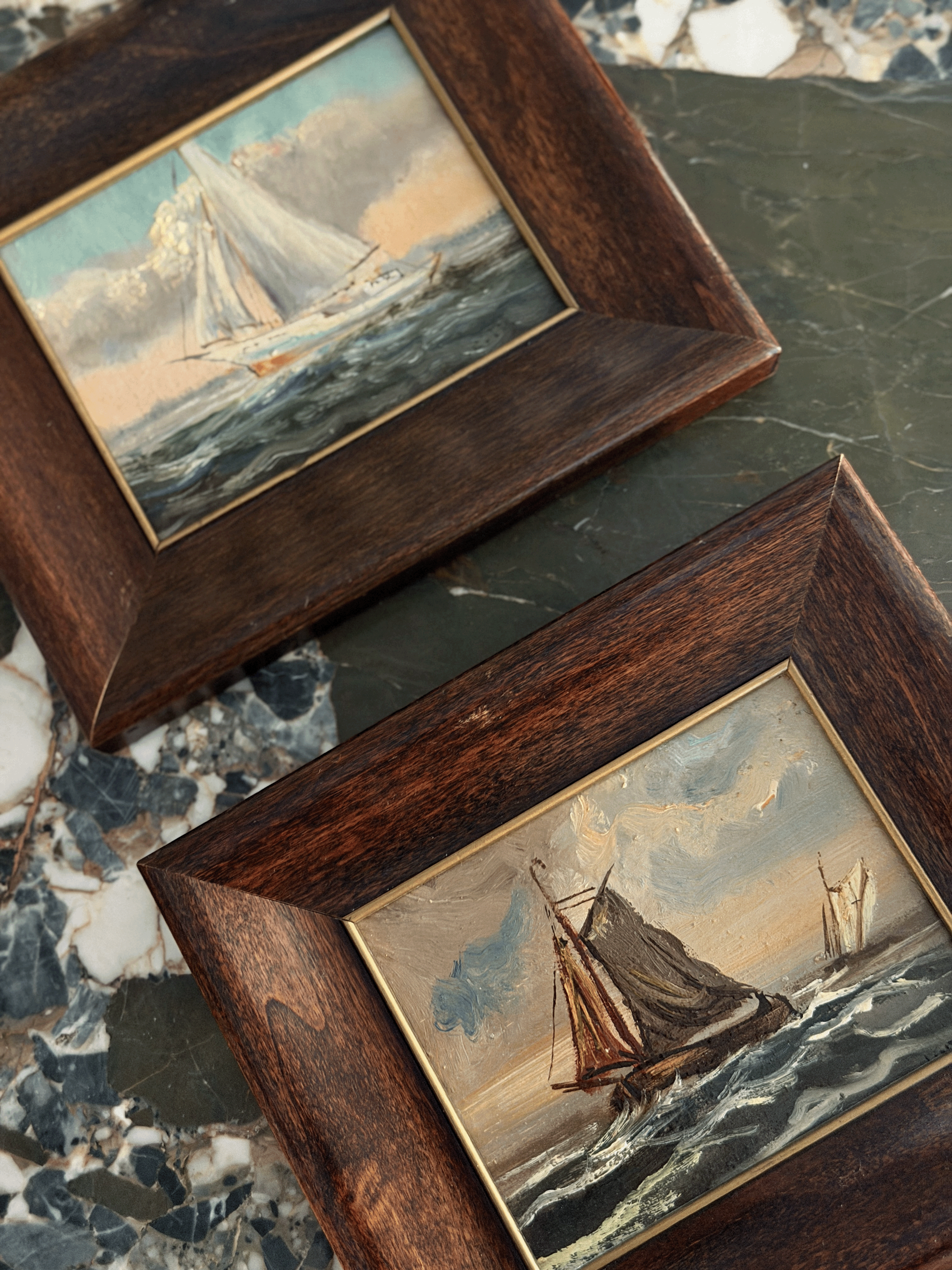 Two framed oil paintings of sailboats on the sea, set in dark brown wood frames with gold inner trim, displayed on a dark marble surface.