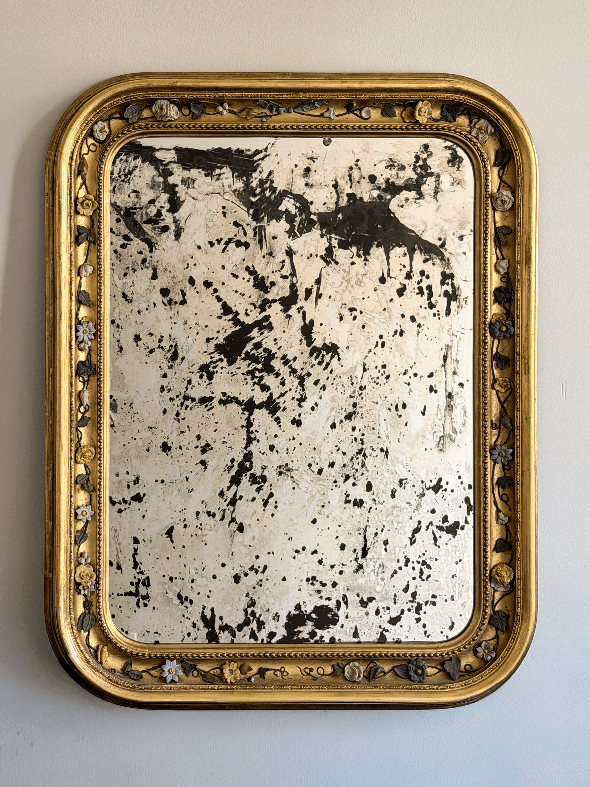 Front view of a 37 by 30 inch 19th-century French Louis Philippe mirror with a gold leaf giltwood frame, rounded top corners, an intricate porcelain floral vine border, and highly distressed, heavily foxed antiqued glass hanging on a light-colored wall.