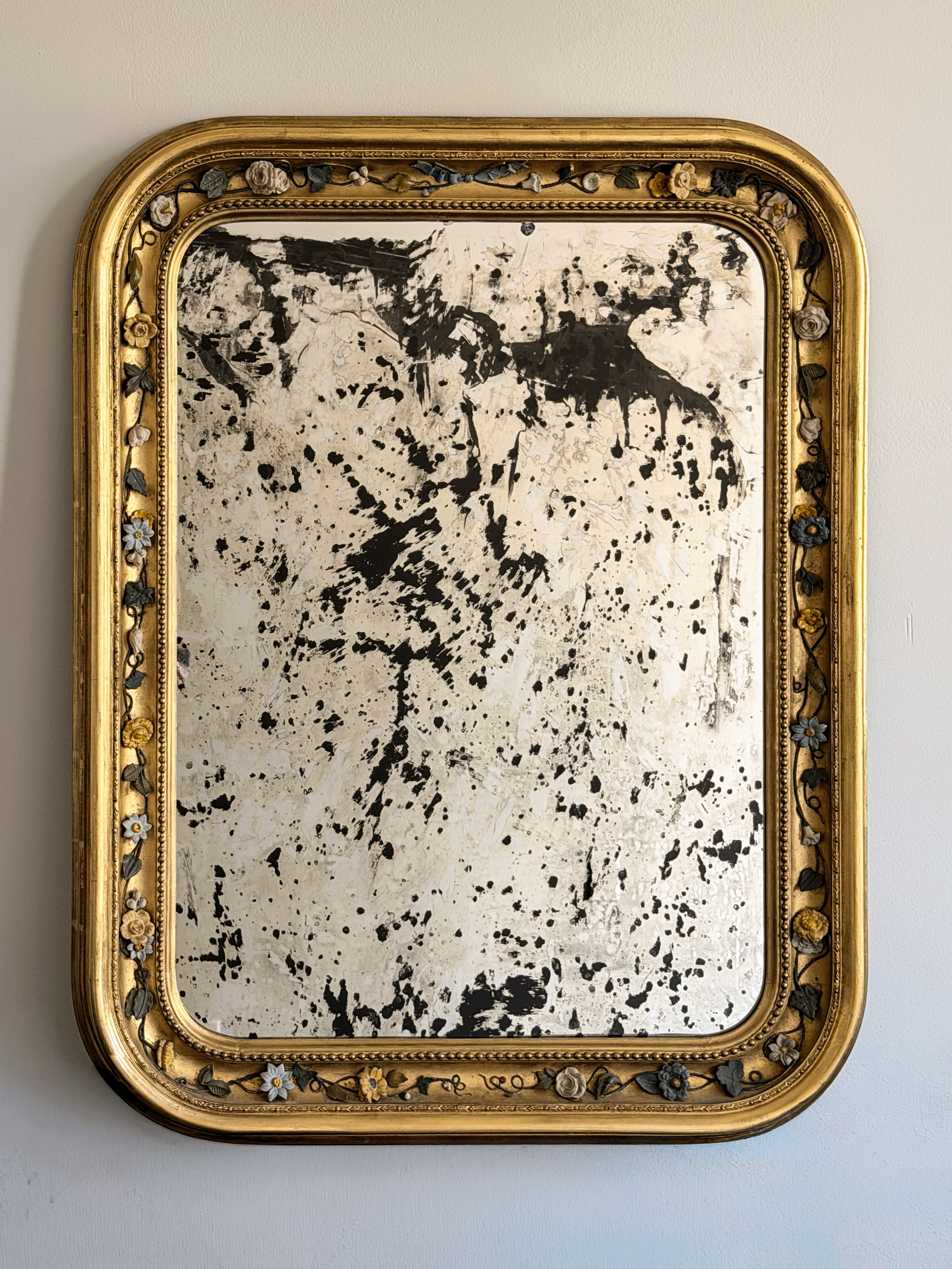 Front view of a 37 by 30 inch 19th-century French Louis Philippe mirror with a gold leaf giltwood frame, rounded top corners, an intricate porcelain floral vine border, and highly distressed, heavily foxed antiqued glass hanging on a light-colored wall.