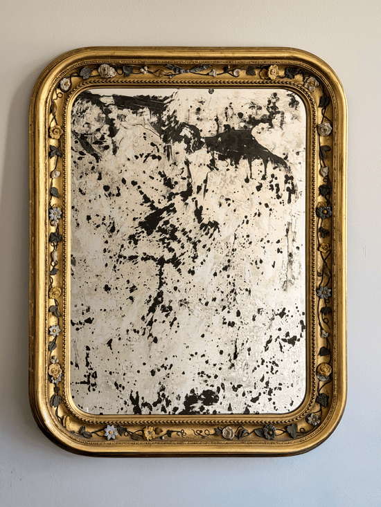 Front view of a 37 by 30 inch 19th-century French Louis Philippe mirror with a gold leaf giltwood frame, rounded top corners, an intricate porcelain floral vine border, and highly distressed, heavily foxed antiqued glass hanging on a light-colored wall.