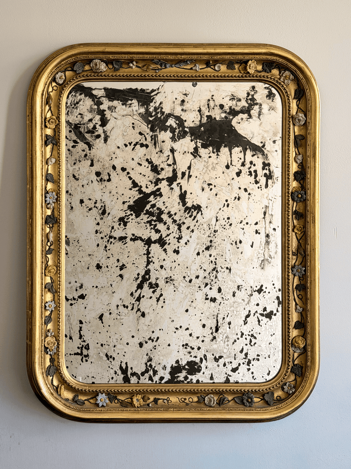 Front view of a 37 by 30 inch 19th-century French Louis Philippe mirror with a gold leaf giltwood frame, rounded top corners, an intricate porcelain floral vine border, and highly distressed, heavily foxed antiqued glass hanging on a light-colored wall.