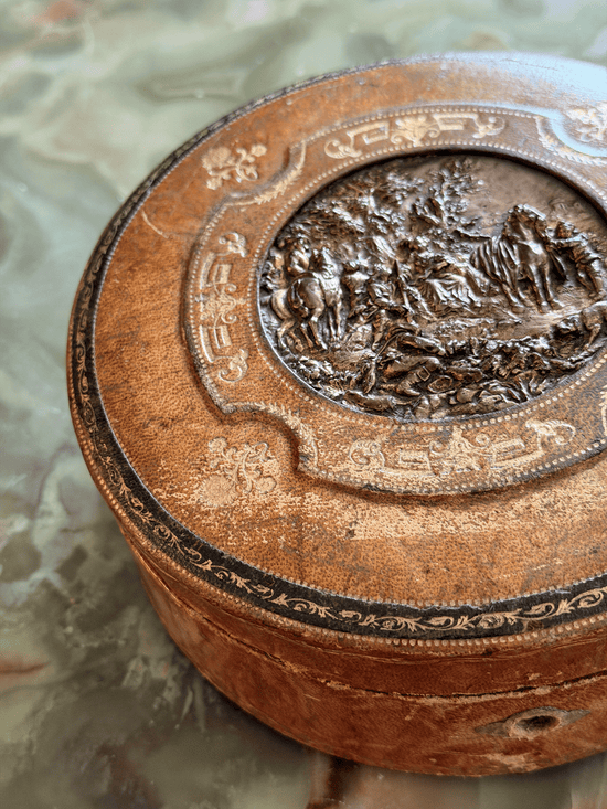 An antique round wooden box with a carved lid featuring a central relief scene and decorative floral patterns, resting on a mottled stone surface.