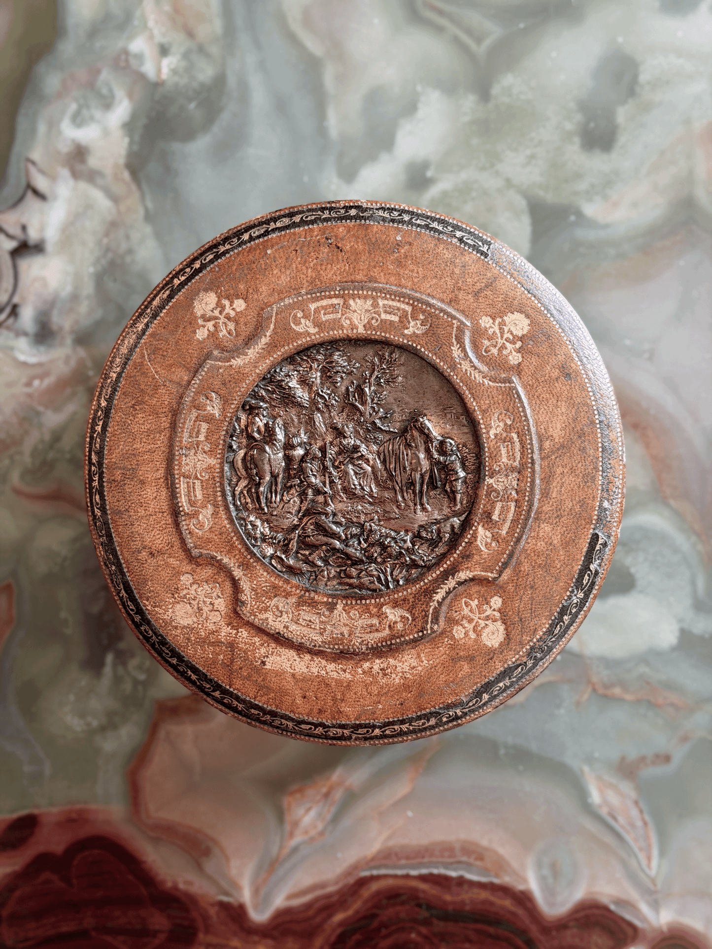 Antique French Grand Tour leather vanity box from the early 20th century, showing a green tooled Morocco leather exterior with a central cast bronze repoussé plaque depicting a classical pastoral garden party scene, opened to reveal a blush-pink ruched silk faille interior and original steel barrel key.